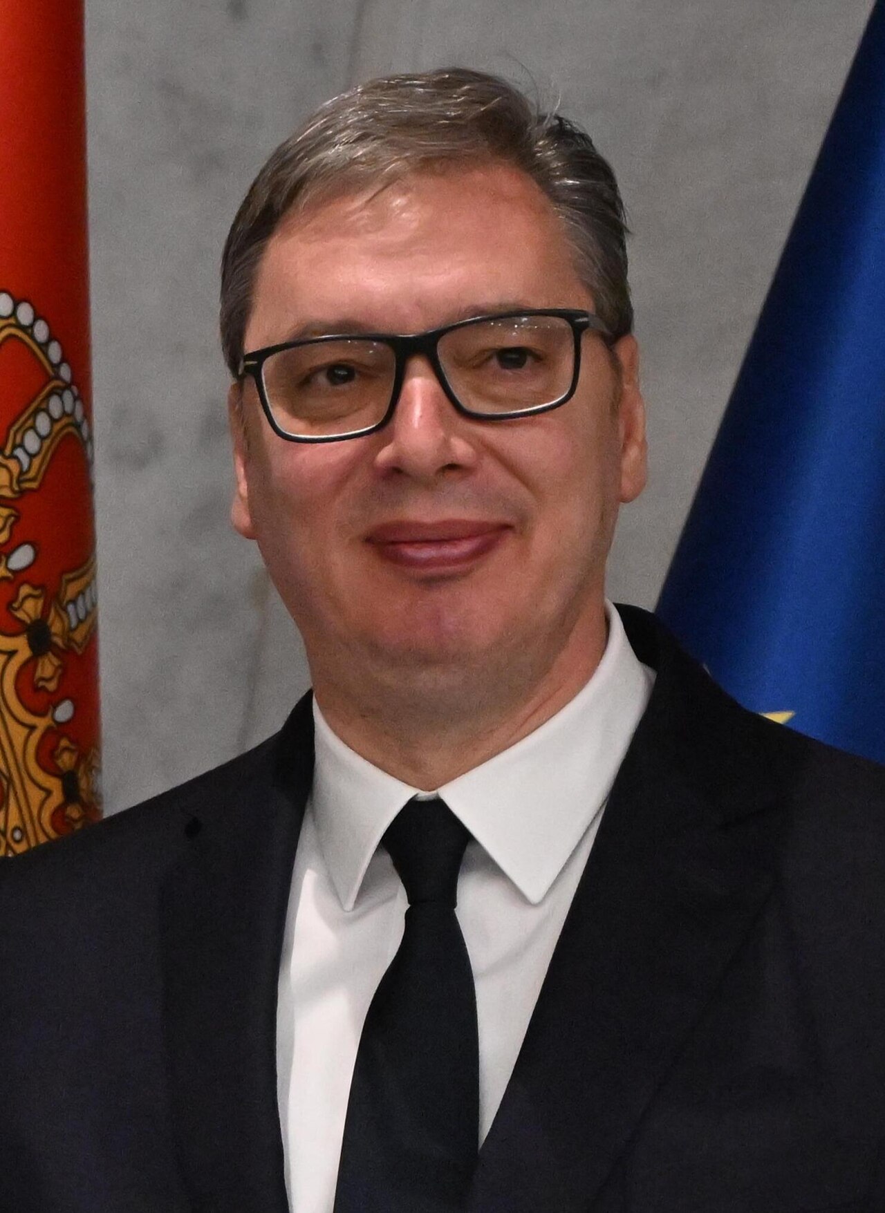 Aleksandar Vučić in glasses, a dark suit and black tie, smiling in front of Serbian and EU flags.