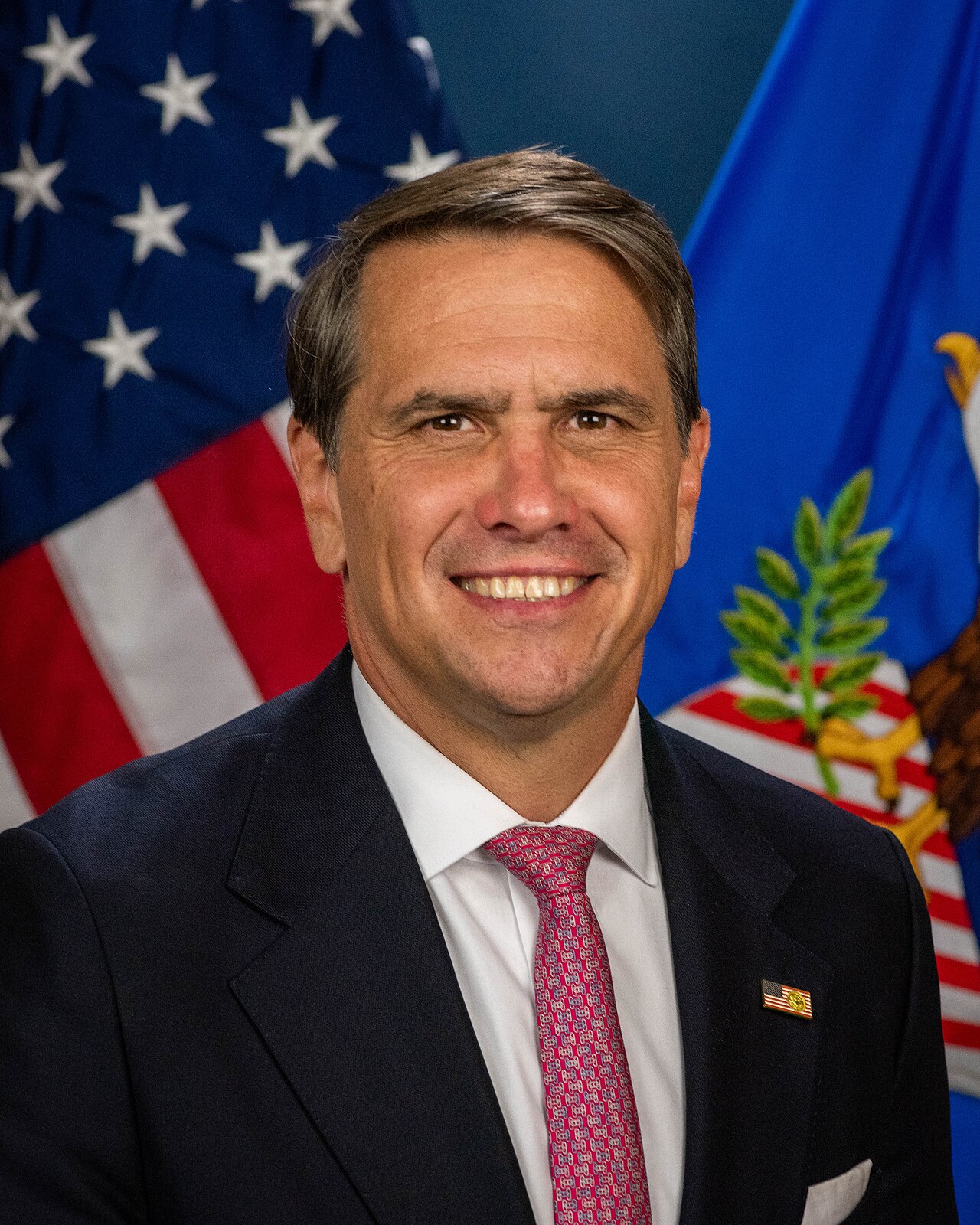A man in a suit with a US flag pin on his lapel, standing in front of American and Department of Justice flags.