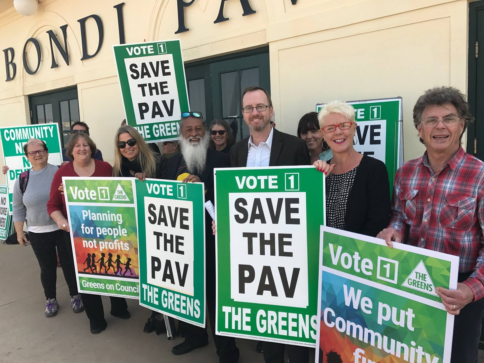 A group of people are holding green signs that say "Vote 1 The Greens" and "Save the PAV". The signs also say "Planning for people not profits" and "We put Community first". The background shows a building with the word "BONDI" visible.