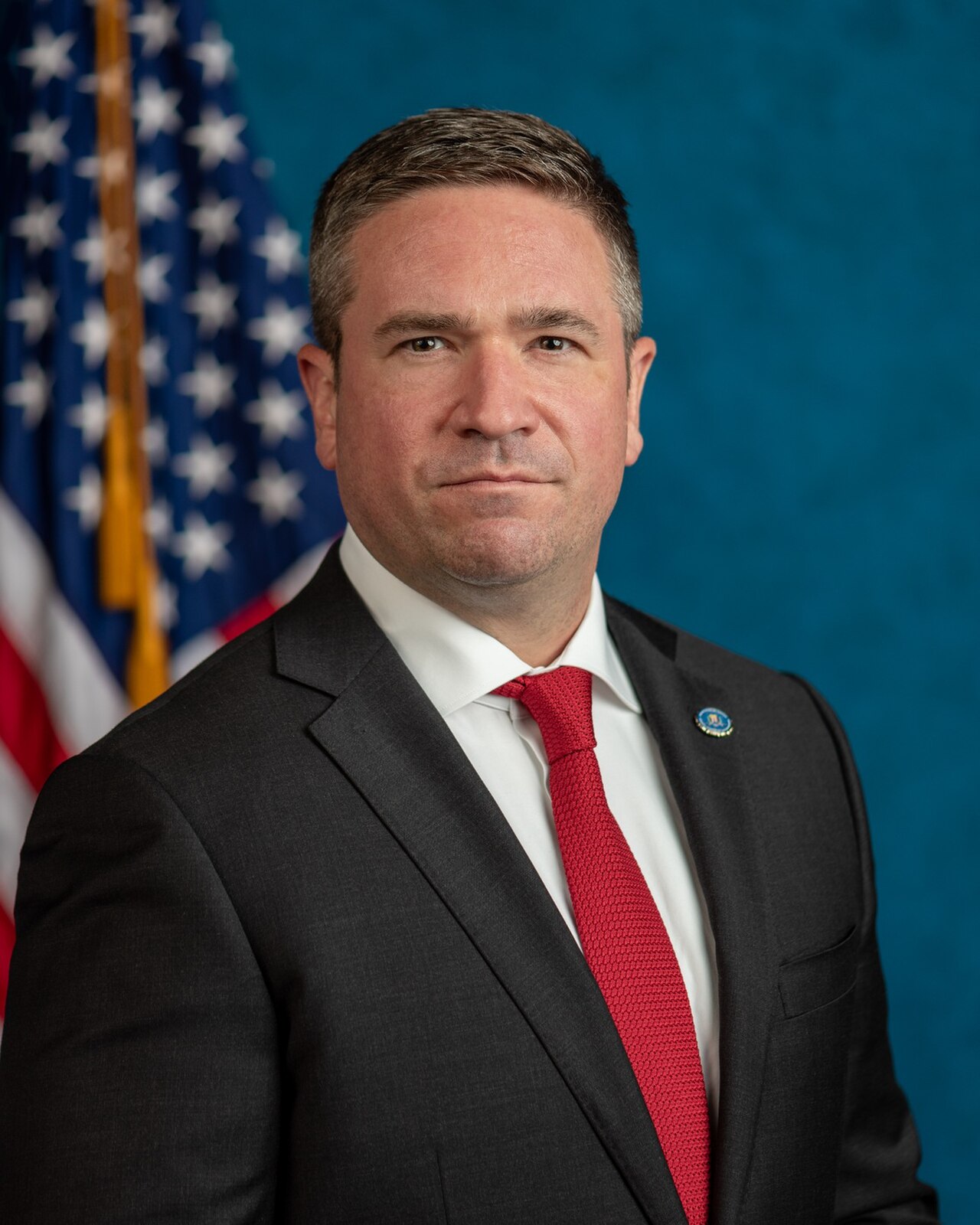 A man in a black suit, white shirt, and red tie, with an American flag behind him.