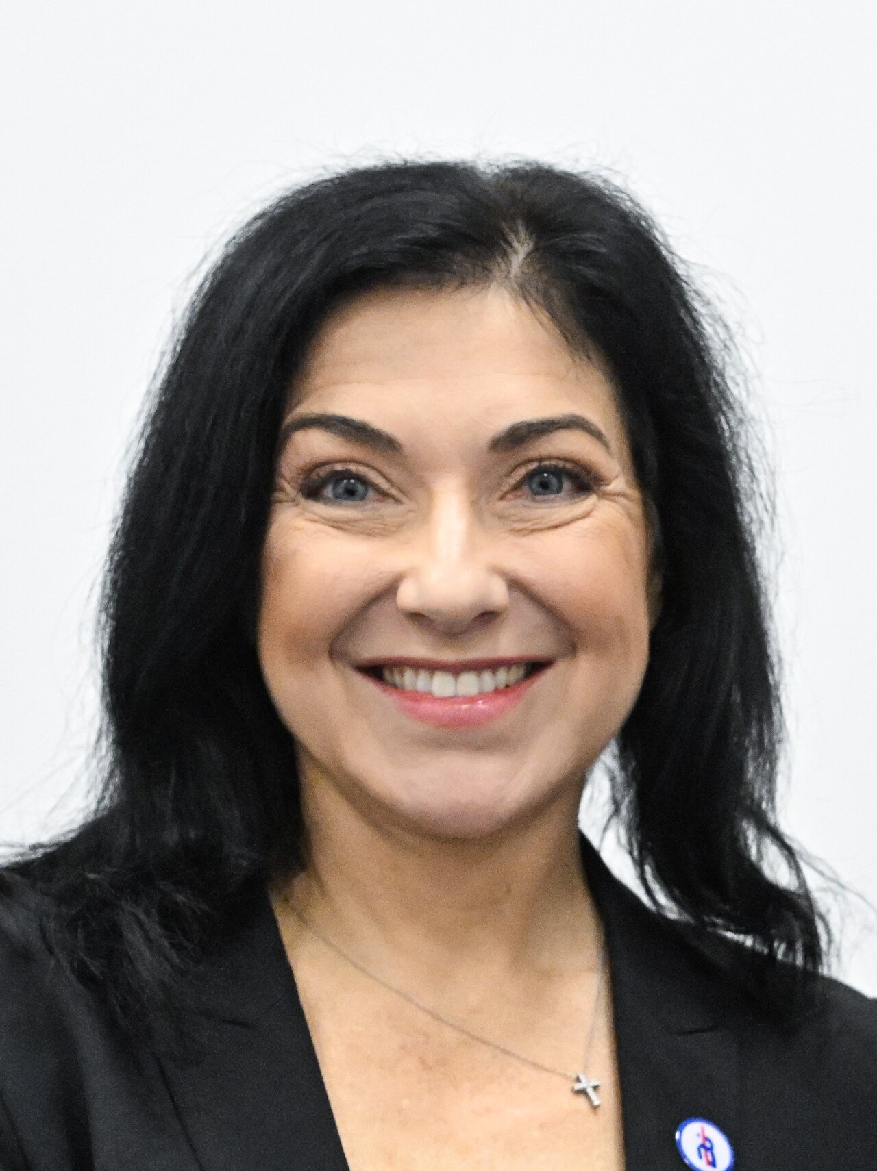 A smiling middle-aged woman with long dark hair wearing a black blazer and a silver necklace with a cross pendant.