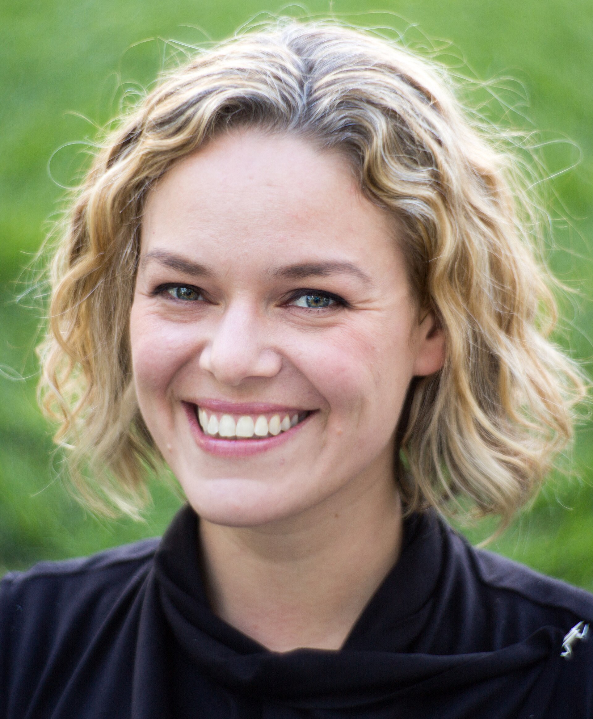 A smiling woman with short, wavy blonde hair and a black top, against a blurred green background.