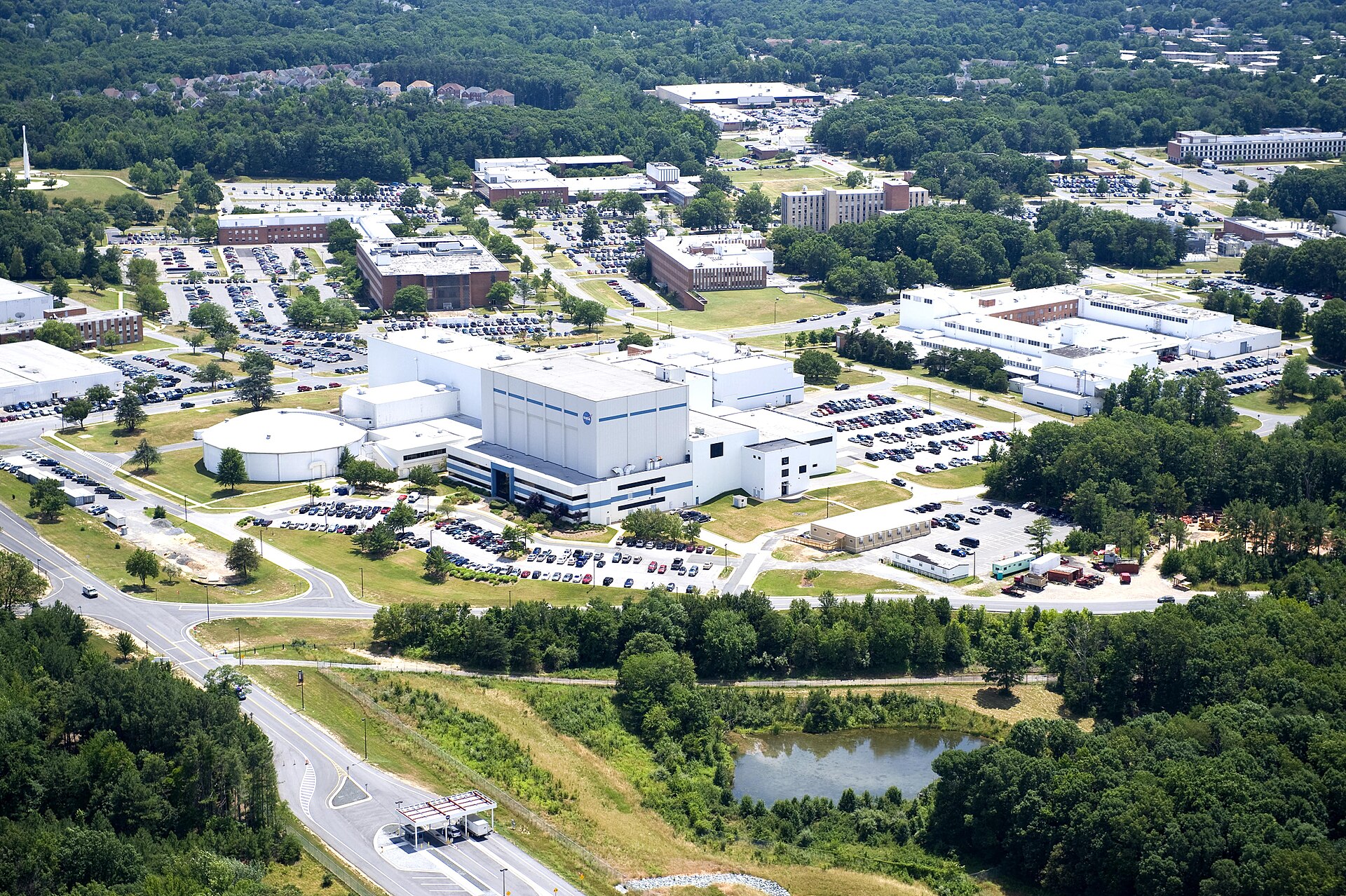 An aerial view of the NASA Goddard Space Flight Center campus, showing multiple large white buildings with blue accents, extensive parking lots filled with cars, and surrounding green trees and landscaping. A prominent building in the foreground displays the NASA logo.