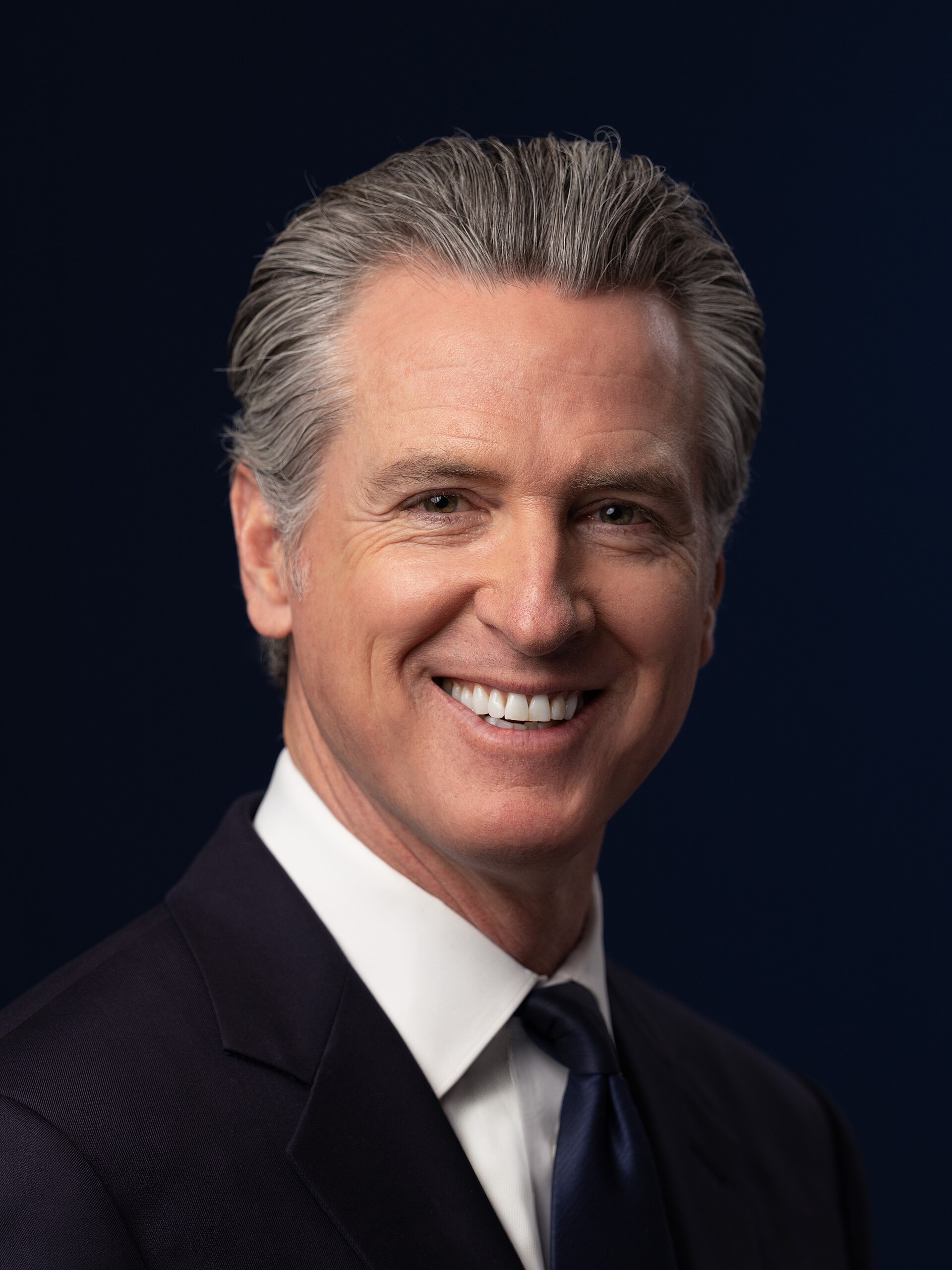 A smiling middle-aged man with graying hair, wearing a dark suit, white shirt, and dark tie, against a dark background.