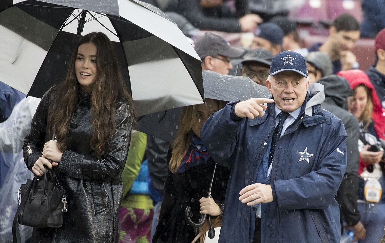 Dallas Cowboys owner Jerry Jones gesturing in the rain before a 2017 game while a woman holds an umbrella nearby.
