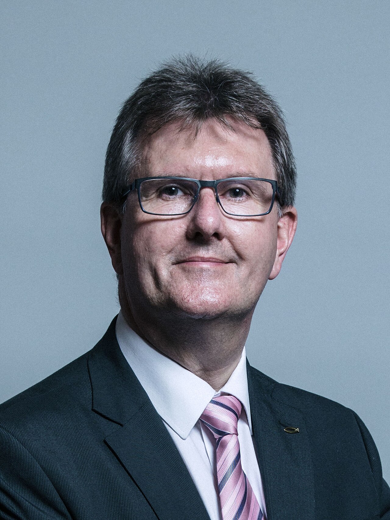 A headshot of Jeffrey Donaldson, a white man in his 60s, wearing glasses, a dark suit, a white shirt, and a pink and blue striped tie.