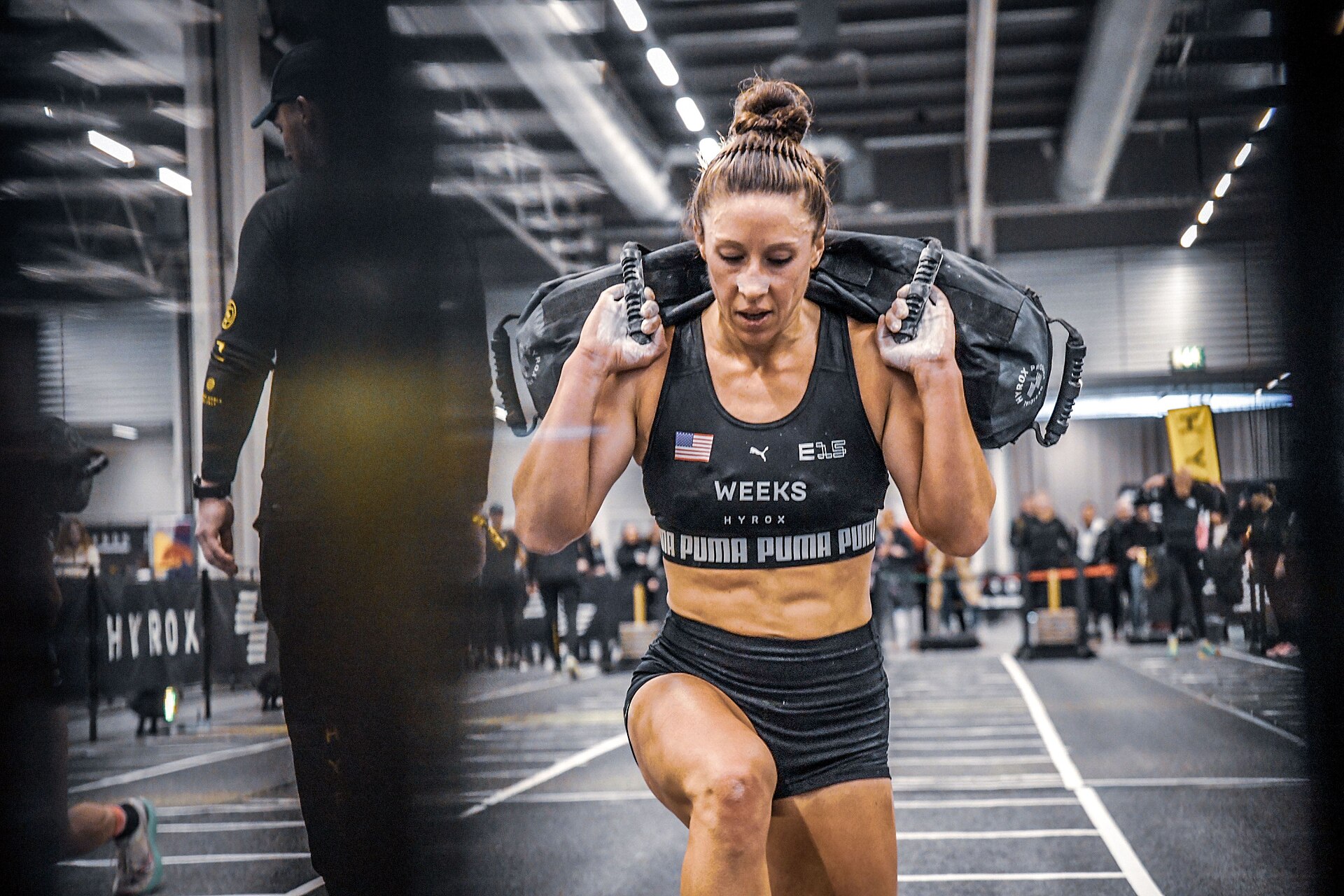 A female athlete with her hair in a bun, wearing a black sports bra and shorts, carries a weighted bag on her shoulders during a Hyrox competition.