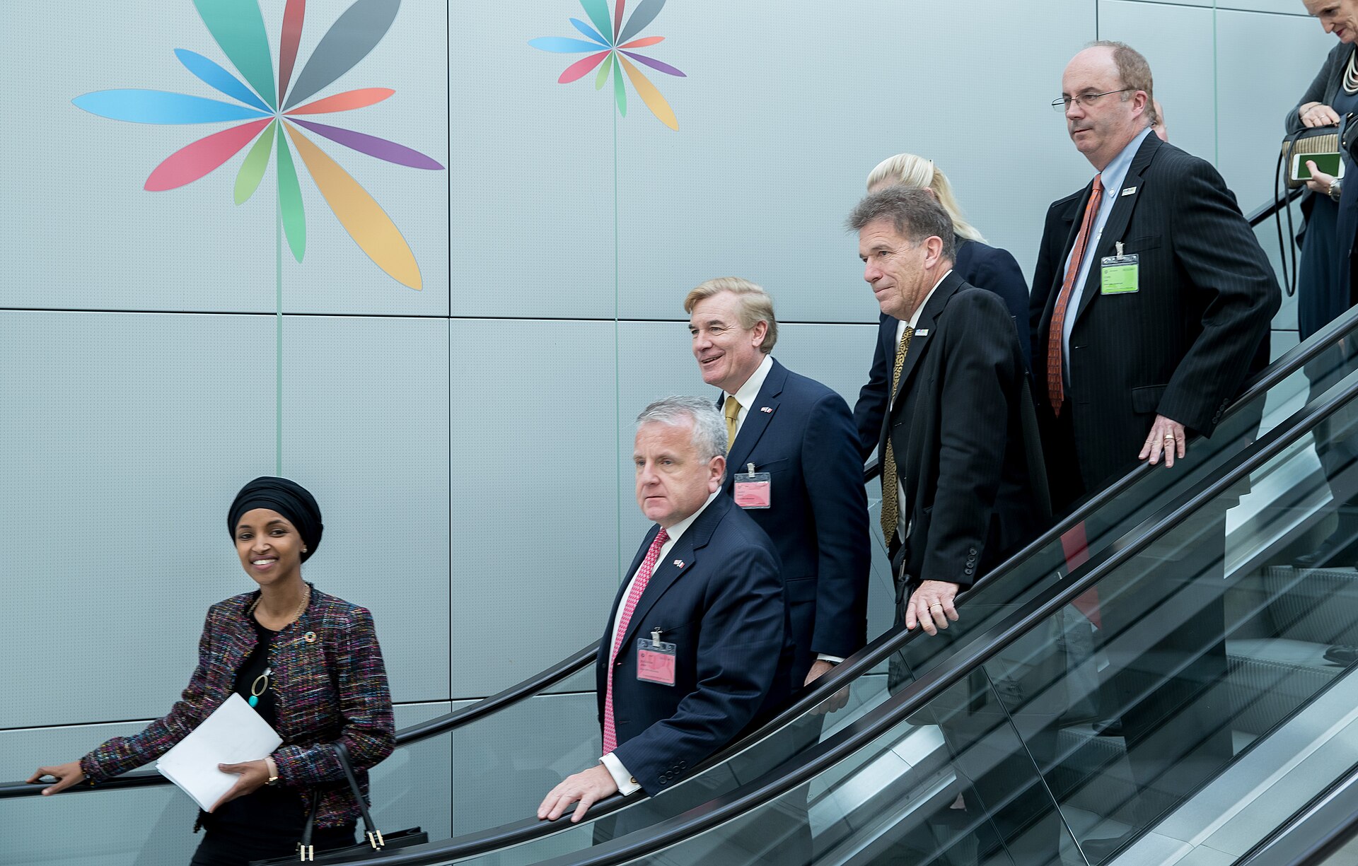 Ilhan Omar, wearing a black hijab and a colorful tweed jacket, smiles as she holds a white folder and a black purse while walking on an escalator with several men in suits.