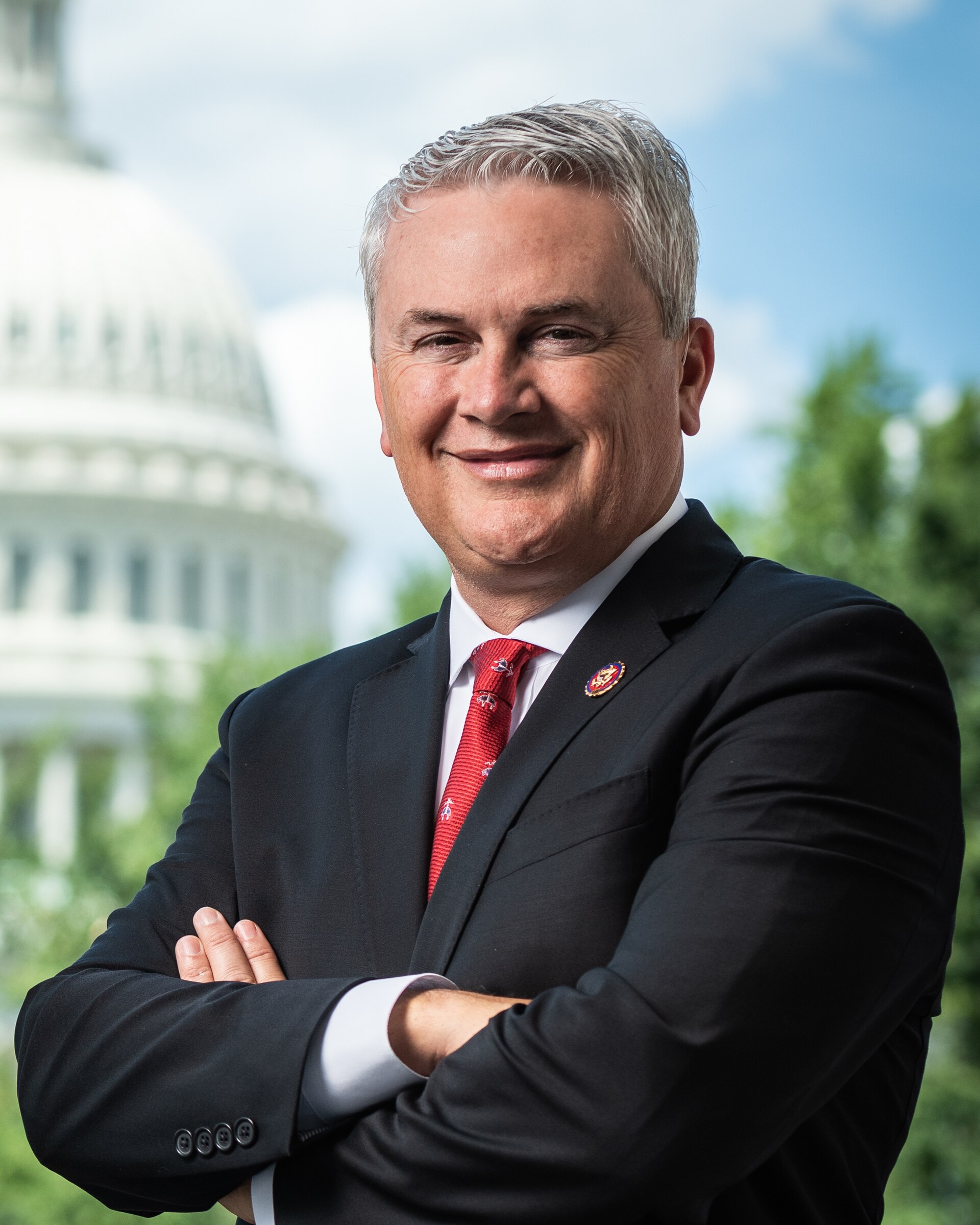 A middle-aged man with short gray hair, wearing a black suit, white shirt, and red tie, with his arms crossed, standing in front of a blurred background of the US Capitol building.
