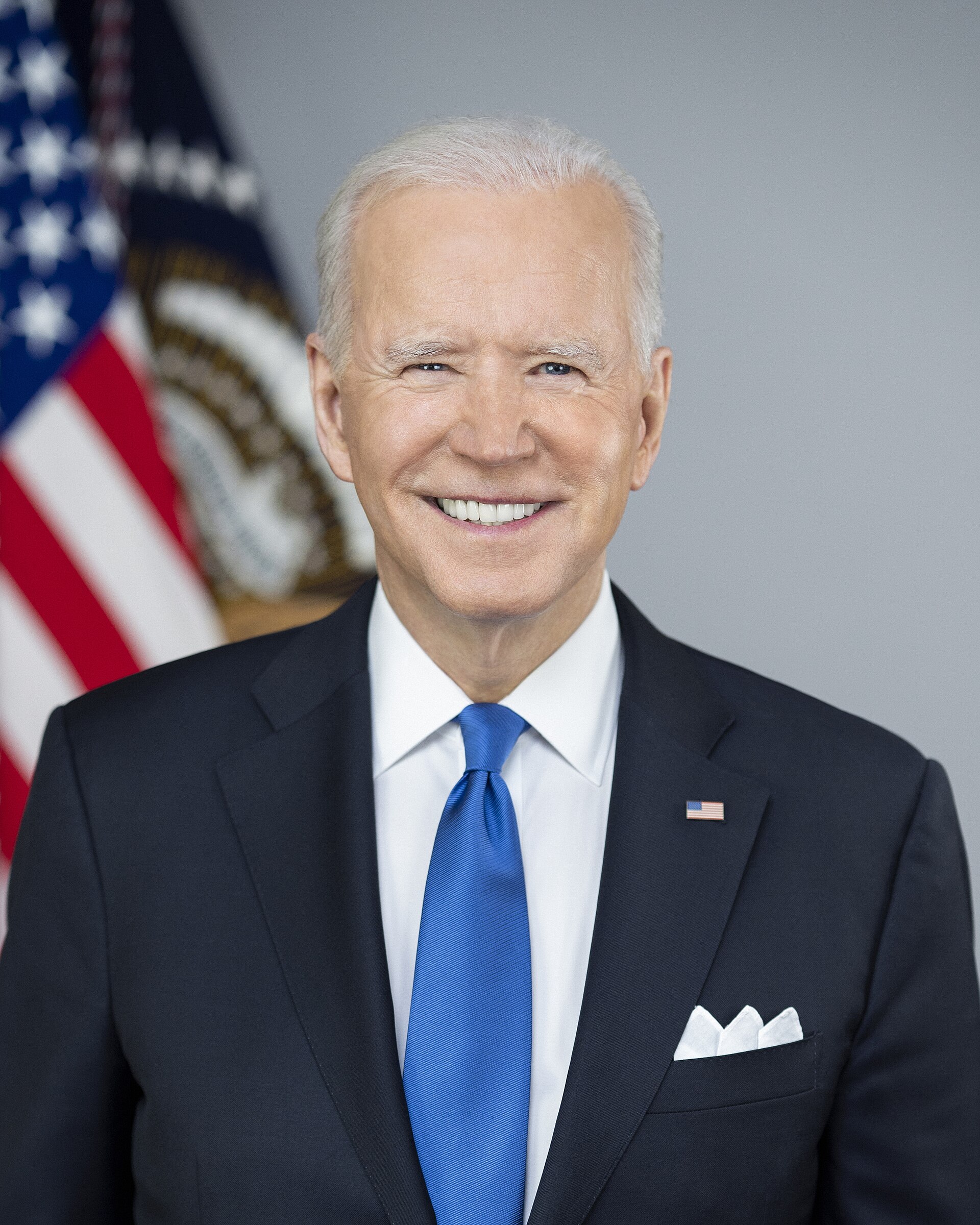 A smiling older white man in a suit with a blue tie and an American flag pin on his lapel, with a blurred American flag and another flag behind him.