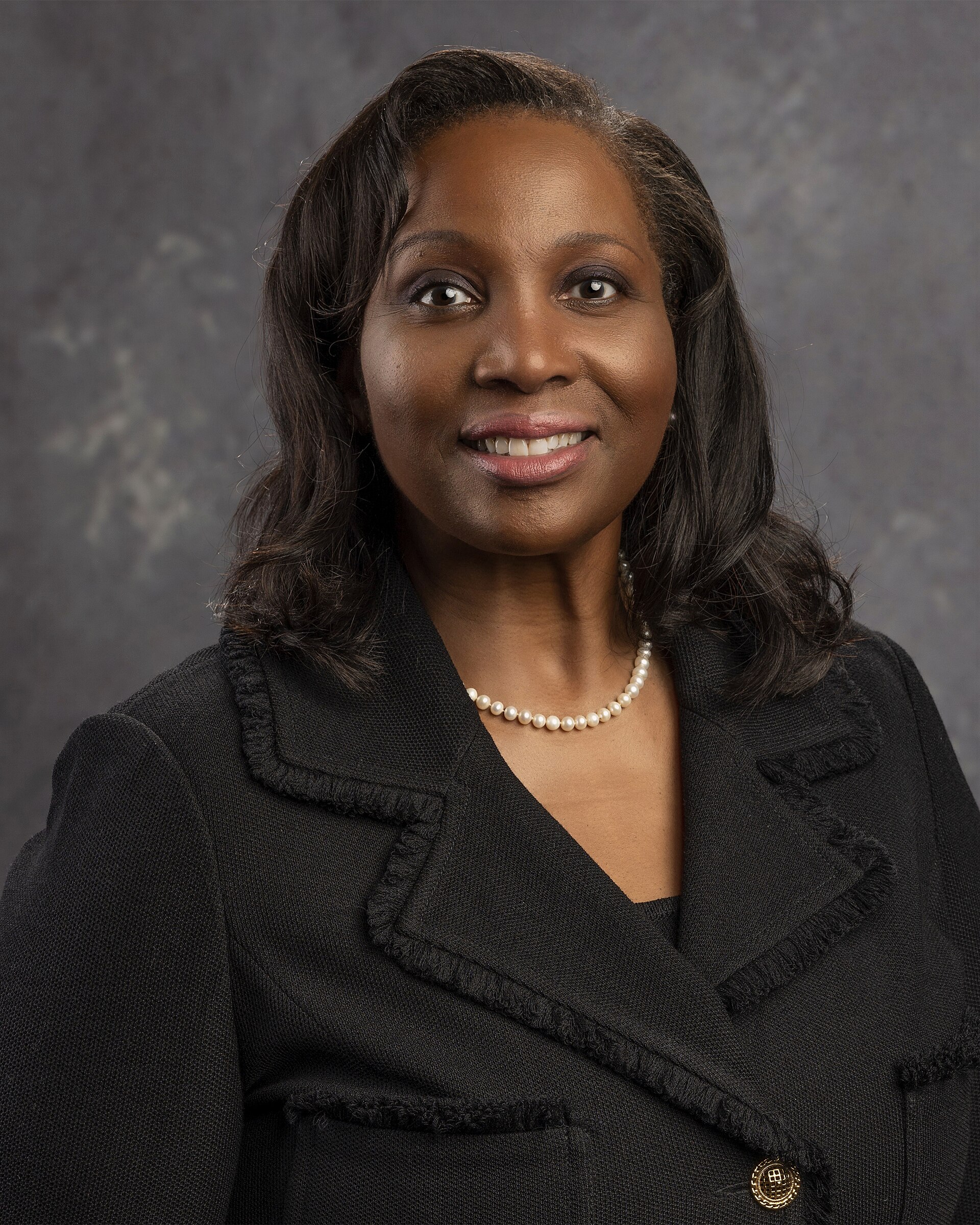 A smiling Black woman with shoulder-length dark hair, wearing a black jacket and pearl necklace, against a gray background.