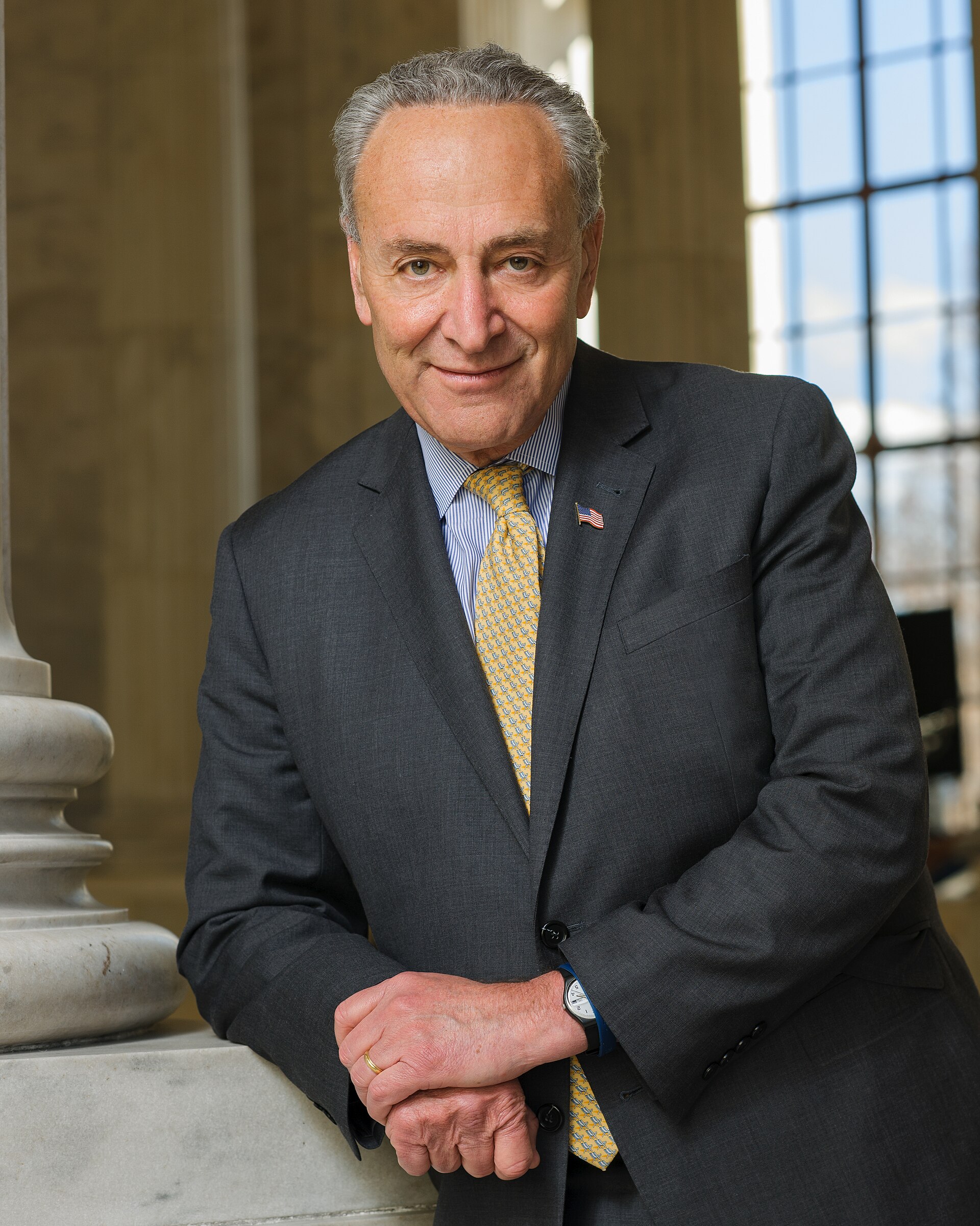 Chuck Schumer, a man with gray hair, wearing a dark suit, a blue and white striped shirt, and a yellow tie with a repeating pattern, leans on a marble pillar and looks towards the camera. He has an American flag lapel pin on his left lapel. His hands are clasped in front of him, and he wears a ring