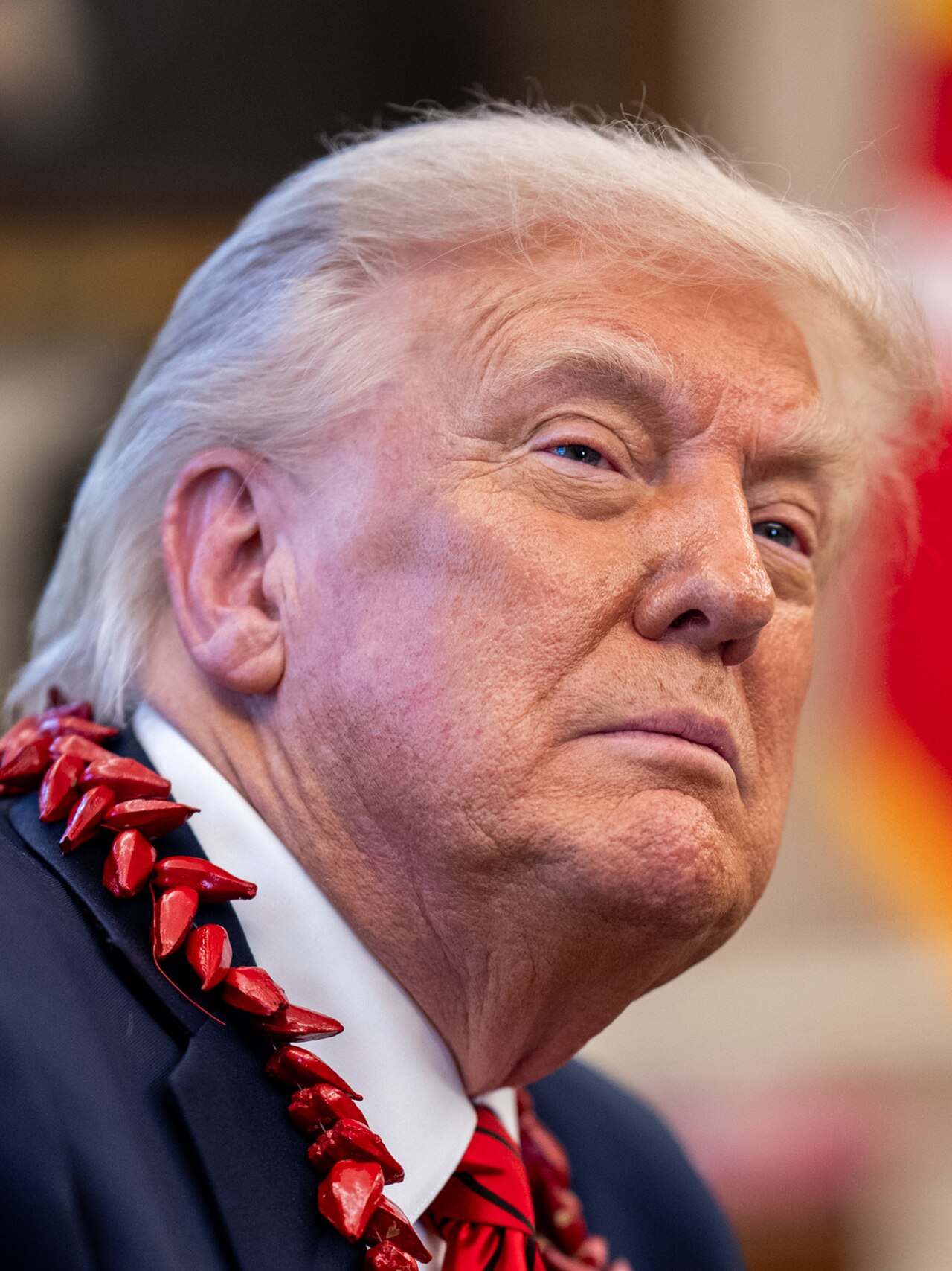 Donald Trump, wearing a dark suit, white shirt, red tie, and a red lei, looking to the right with a serious expression.