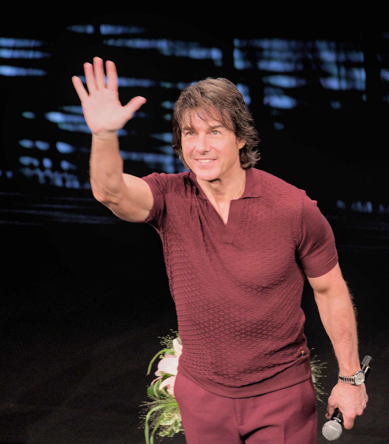 A smiling man with short brown hair, wearing a maroon shirt and pants, waving with his right hand and holding a microphone in his left hand.