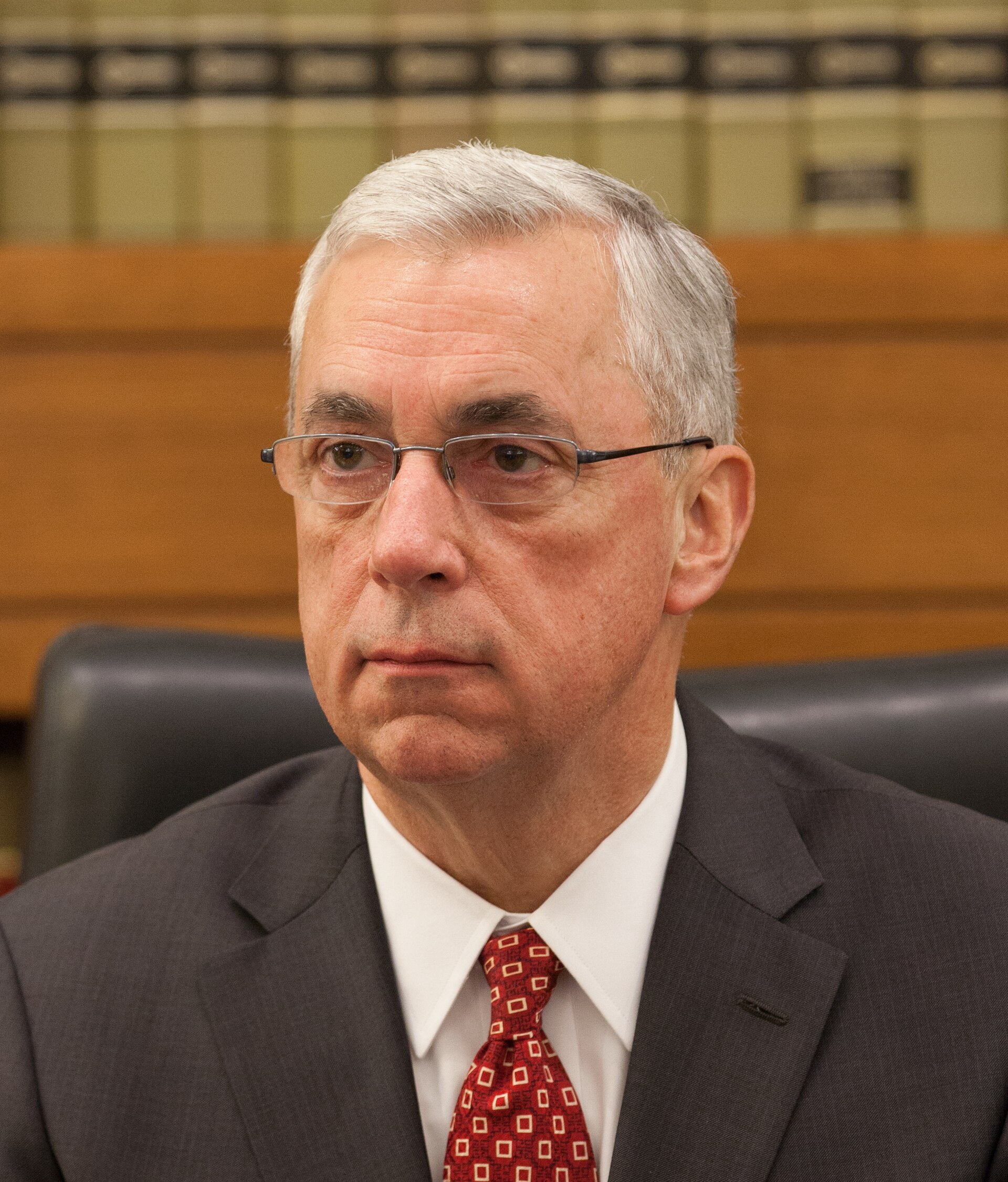 A middle-aged man with short gray hair, wearing glasses, a dark suit, white shirt, and red tie with a pattern.