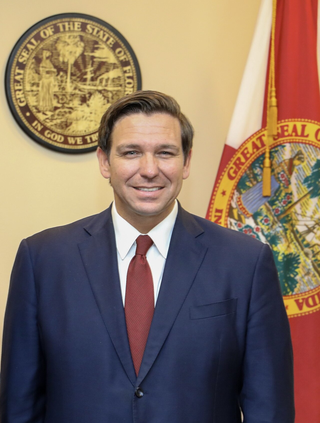 A man in a blue suit and red tie stands in front of a Florida state flag and seal.