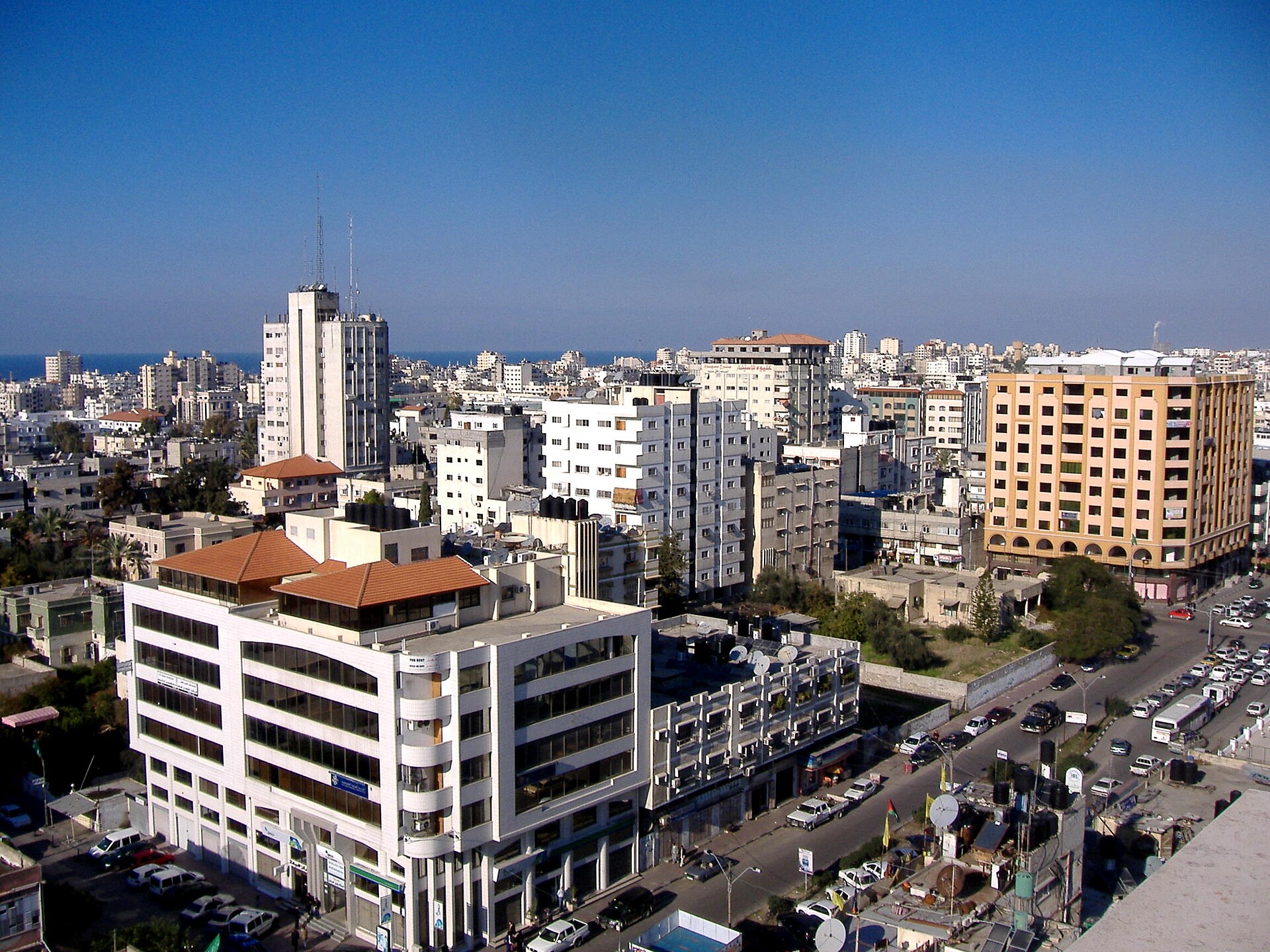 An aerial view of a city skyline with numerous buildings of varying heights and architectural styles under a clear blue sky. In the foreground, a white modern building with large windows and a flat roof is prominent. Further back, a tall, slender white building with antennas on top stands out. To th