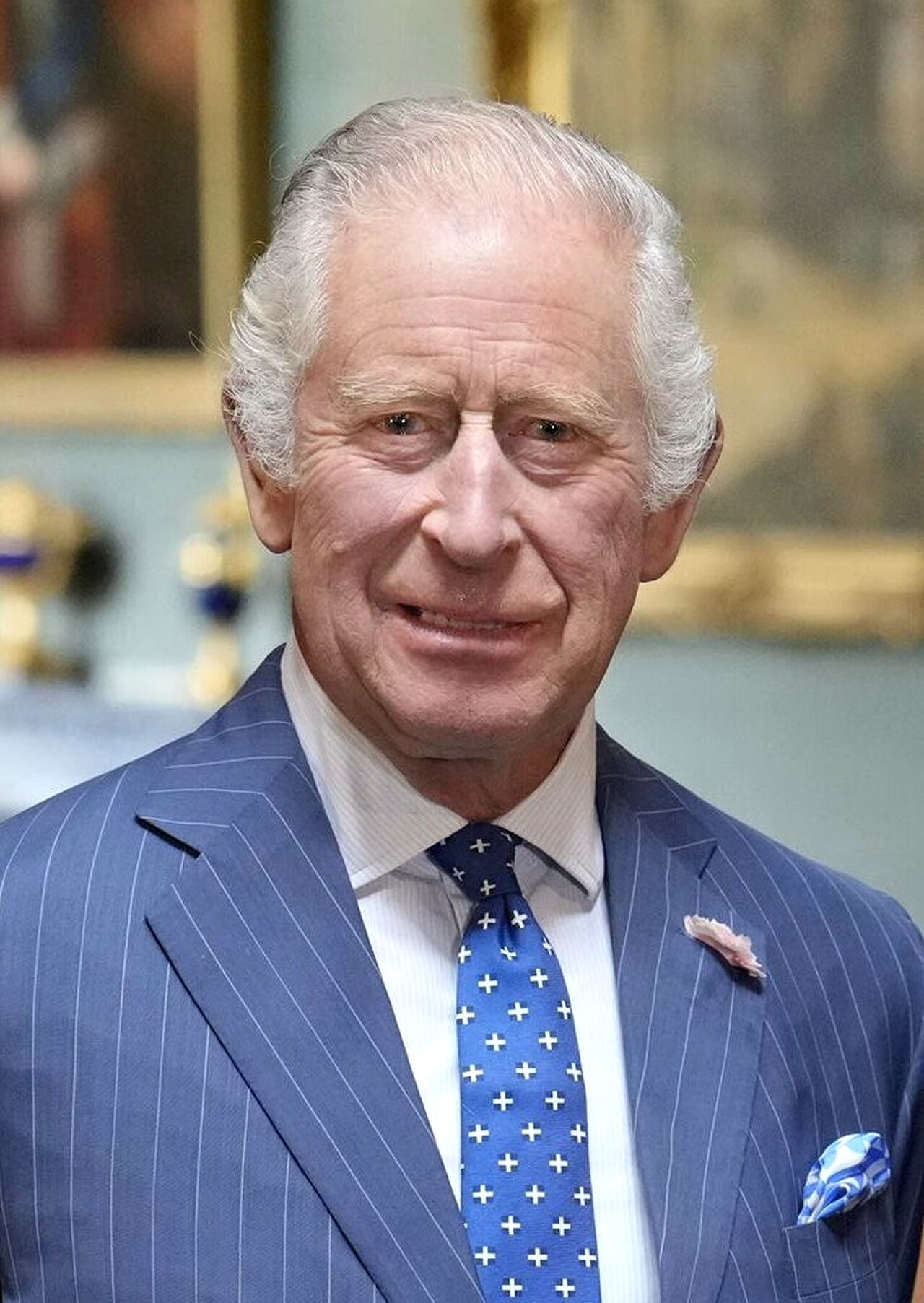 King Charles III, an older man with short white hair, wearing a blue pinstripe suit, white shirt, blue tie with white crosses, and blue pocket square, looking at the camera.