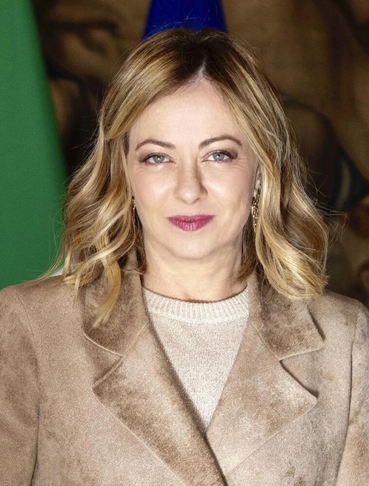 Giorgia Meloni, Prime Minister of Italy, wearing a beige coat and sweater, looking directly at the camera.