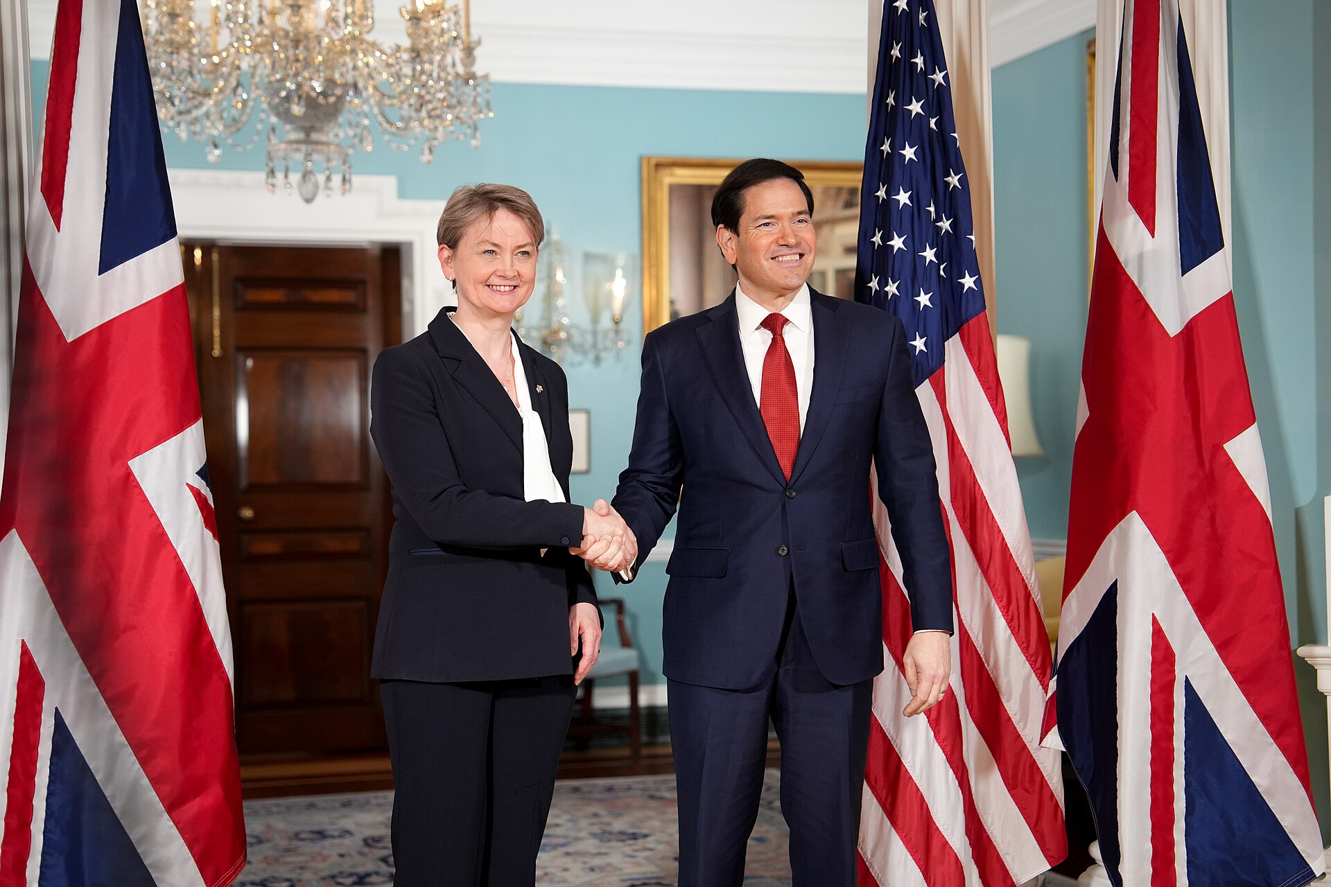 A woman in a dark suit and a man in a dark suit with a red tie shake hands in front of the Union Jack and American flags.