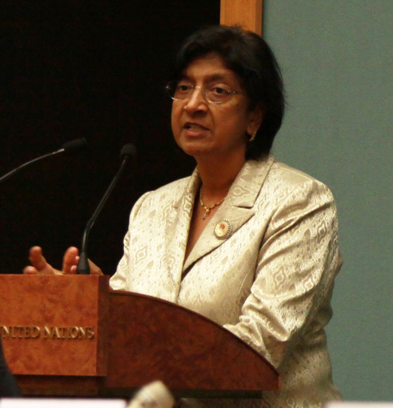 woman with short dark hair, wearing glasses, a cream-colored suit, and a gold necklace, standing at a wooden podium with two microphones, the podium has 'UNITED NATIONS' written on it in gold letters