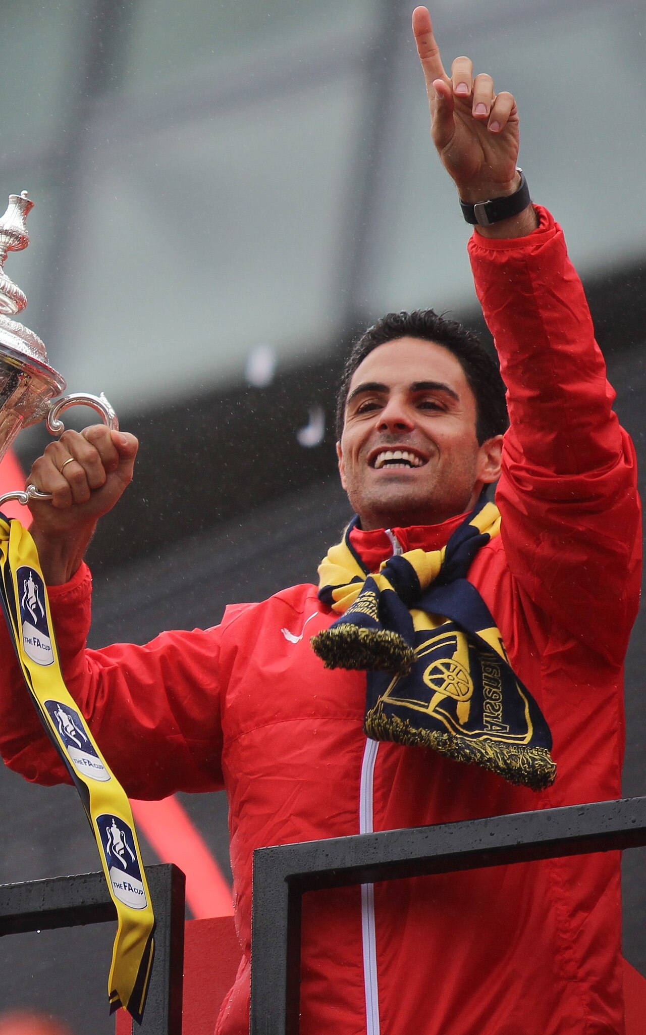 A man with short dark hair wearing a red jacket and a yellow and blue scarf, holding a trophy and pointing upwards with his right hand.