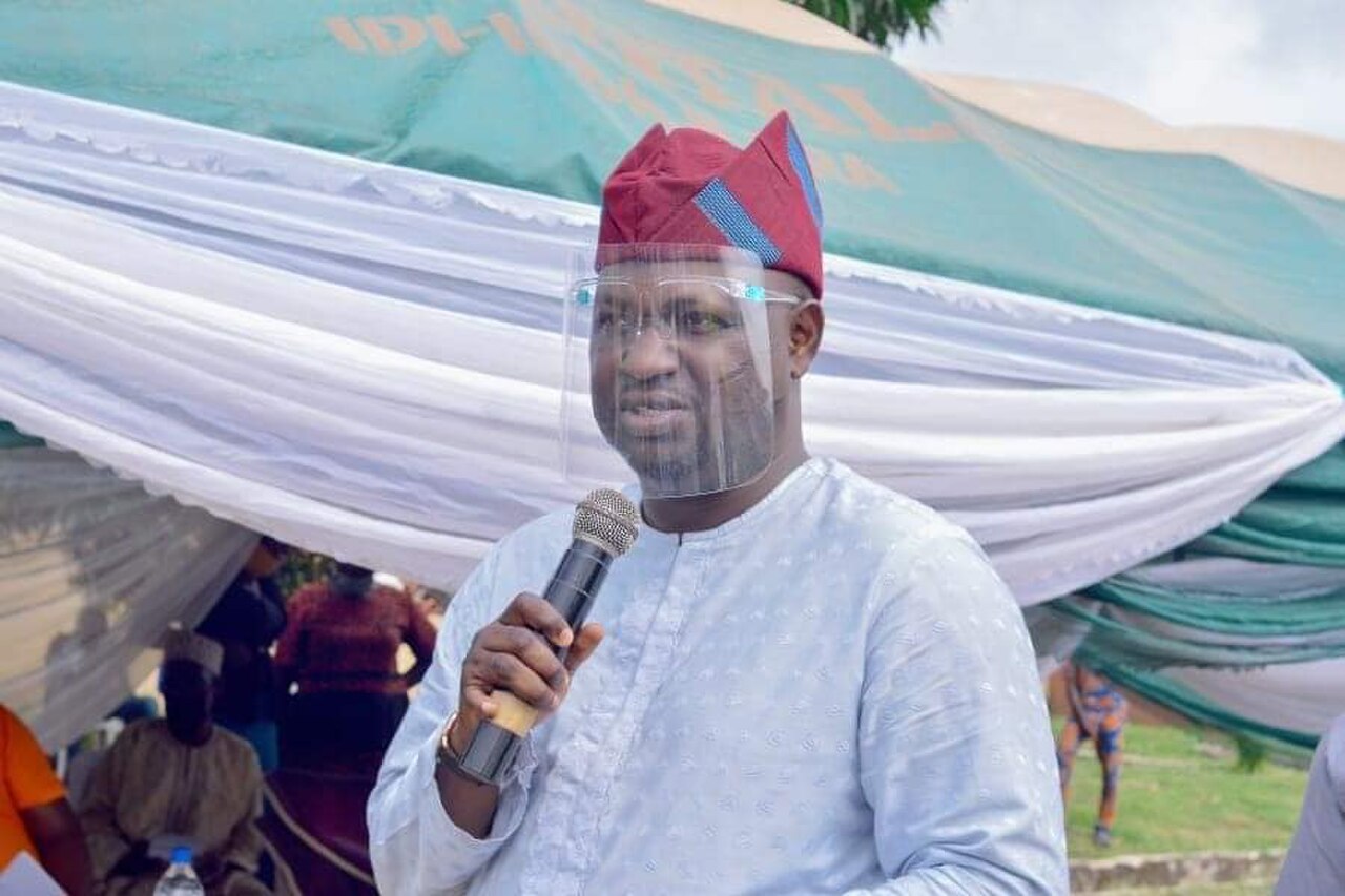 Adeniyi Adeyemi, a man in his 30s, wearing a red cap, a face shield, and a light blue traditional outfit, holding a microphone.