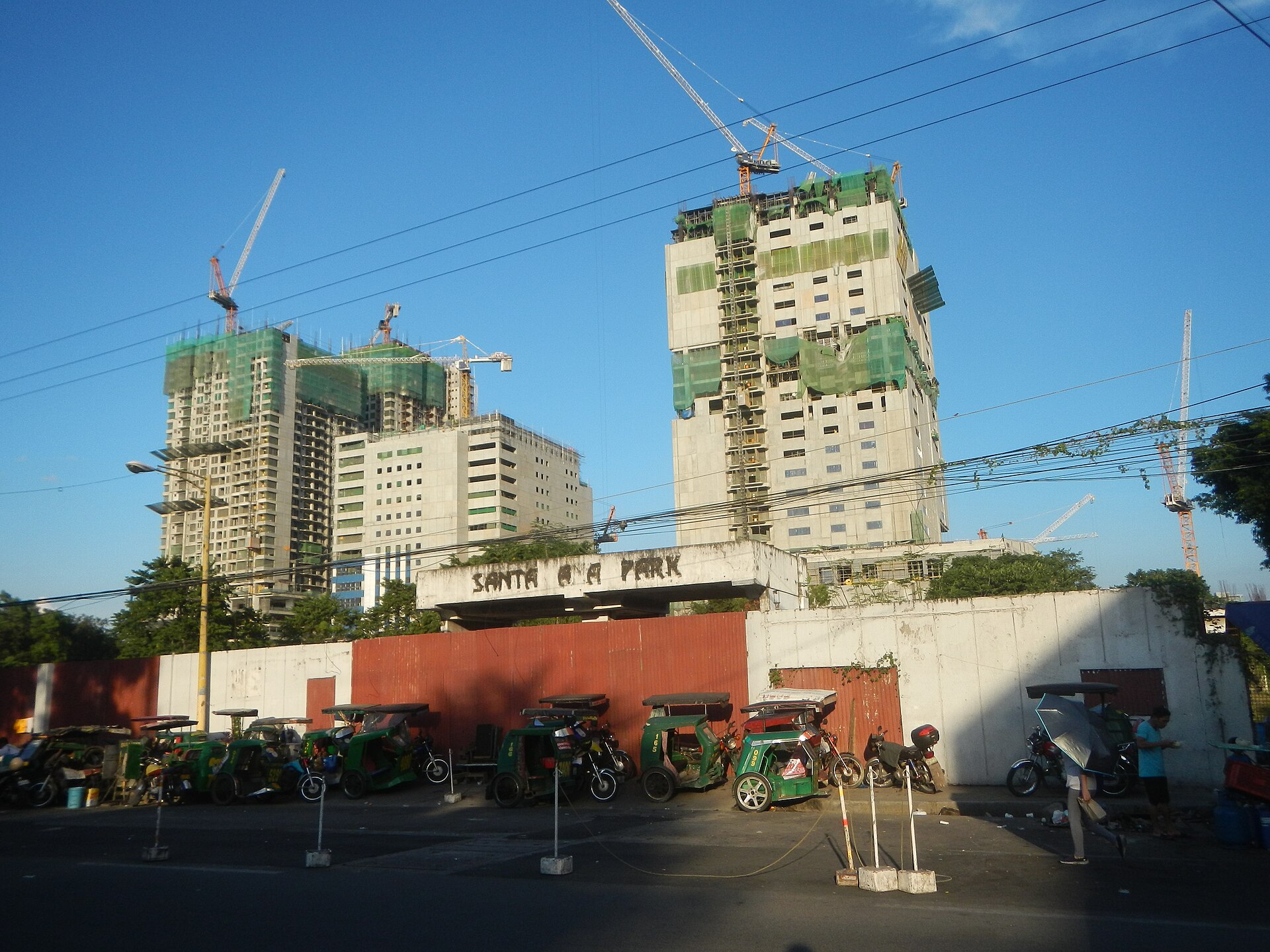 Construction cranes tower over unfinished buildings, with a row of parked auto rickshaws in the foreground.
