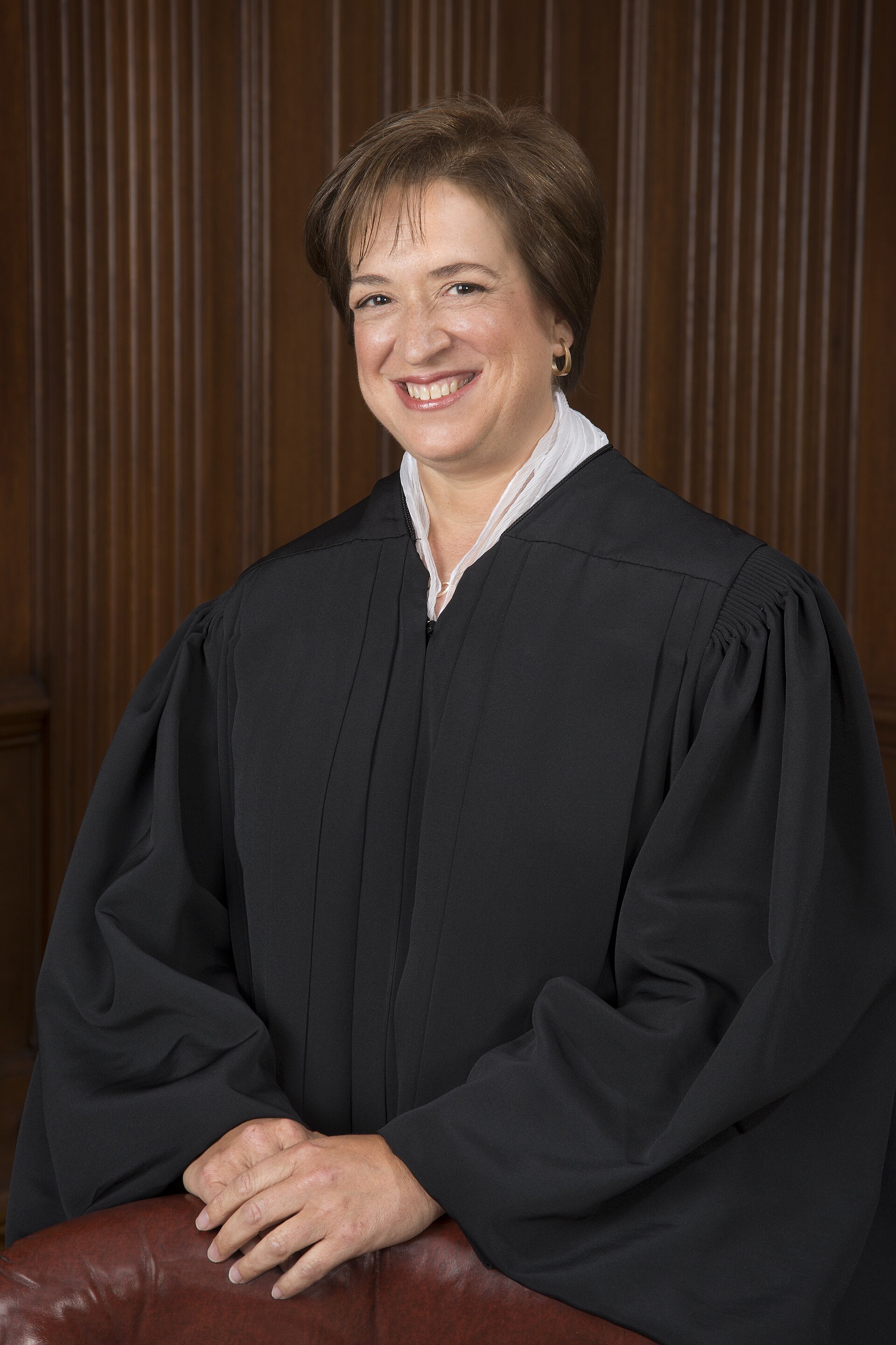Elena Kagan, smiling, in a black judicial robe with a white collared shirt underneath, leaning on a brown leather chair in front of a wood-paneled wall.