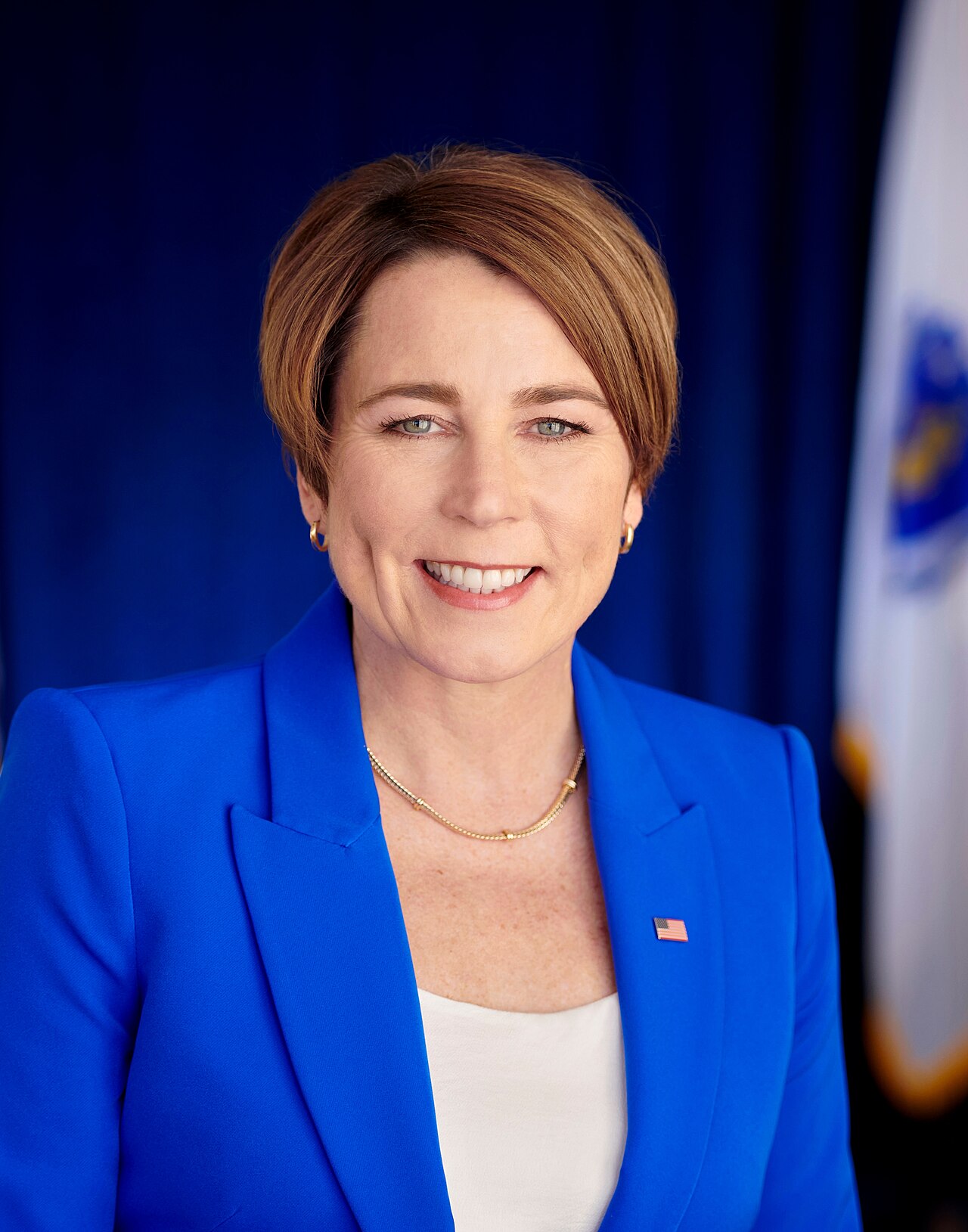 A woman with short brown hair, wearing a blue blazer and a white top, with a gold necklace and earrings, and a small American flag pin on her lapel, smiling and looking at the camera.
