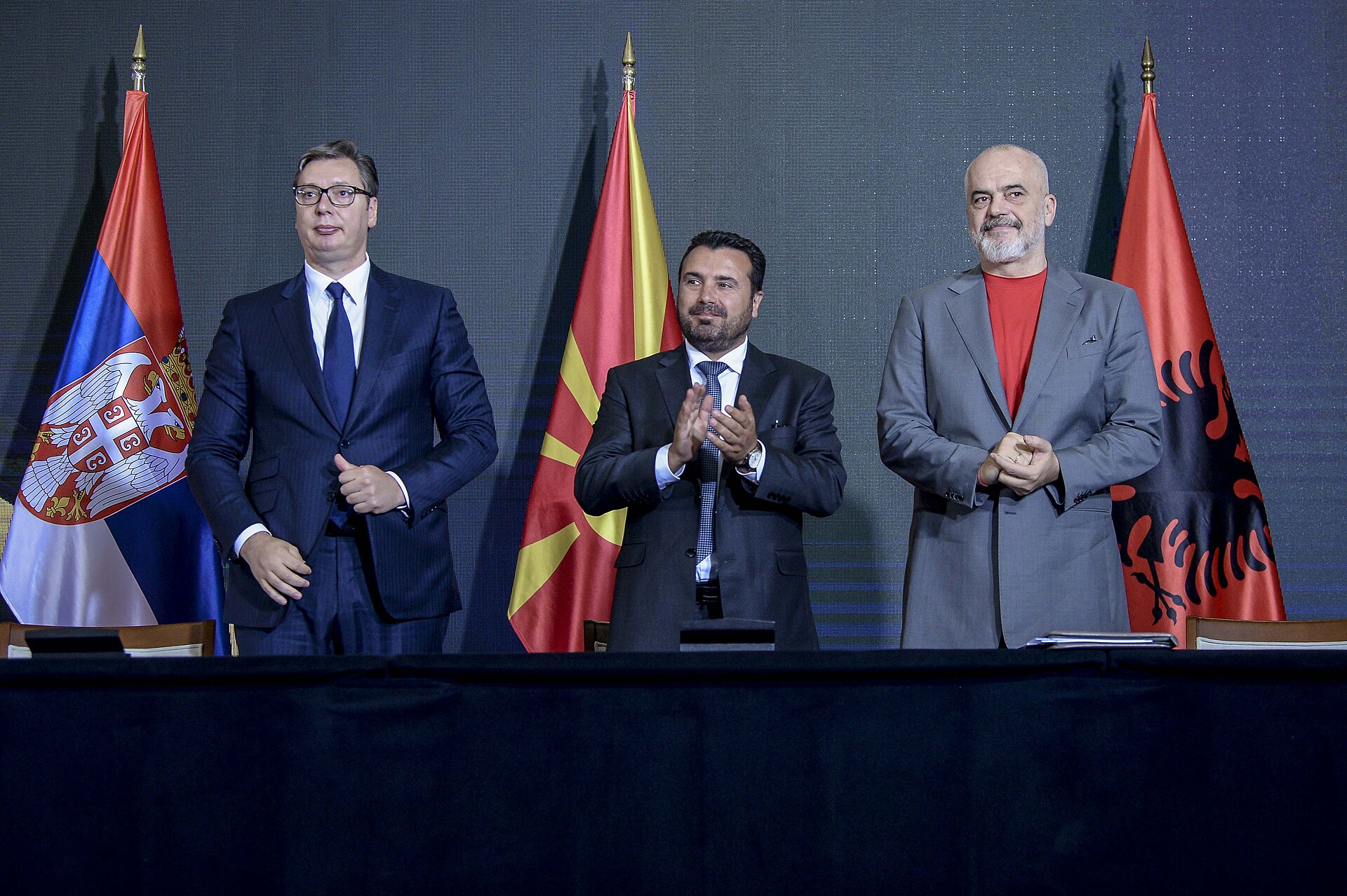 Edi Rama, Prime Minister of Albania, stands between the flags of Serbia and North Macedonia, wearing a grey suit over a red t-shirt.