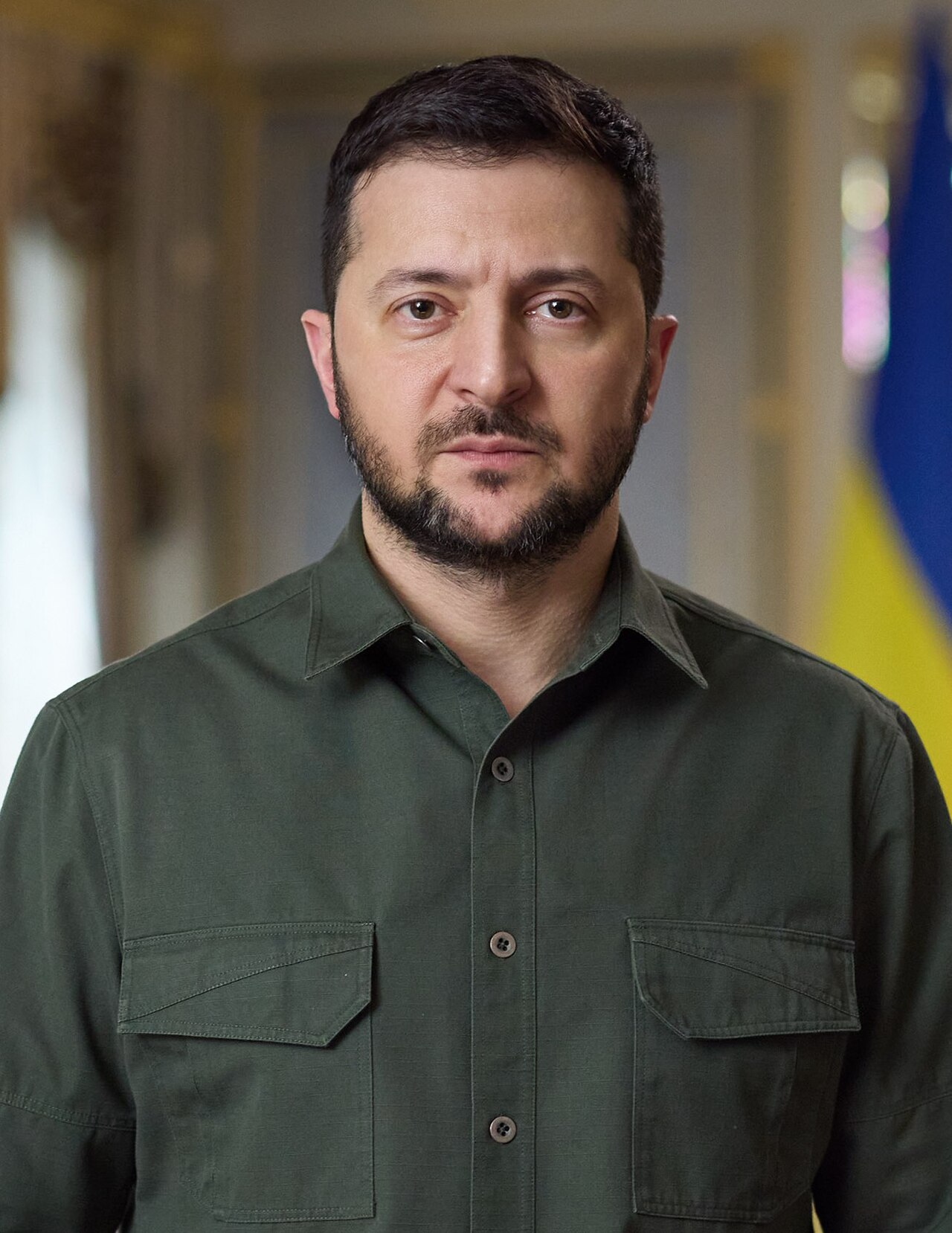 A headshot of Volodymyr Zelensky, President of Ukraine, wearing a dark green button-up shirt. He has a beard and short dark hair. Behind him, a blurred Ukrainian flag is visible.