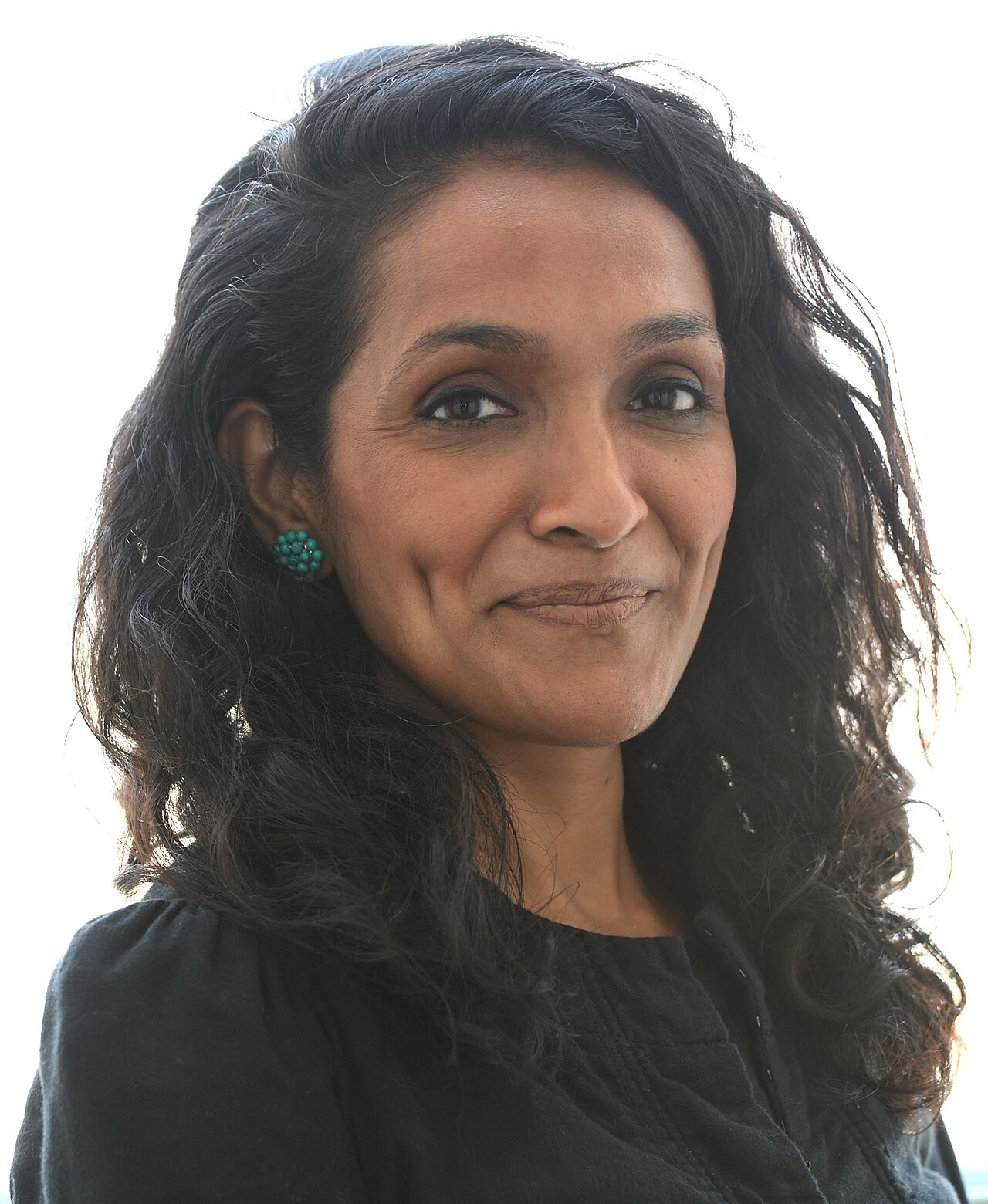 A smiling woman with long, dark, curly hair and green earrings, wearing a black top, against a white background.