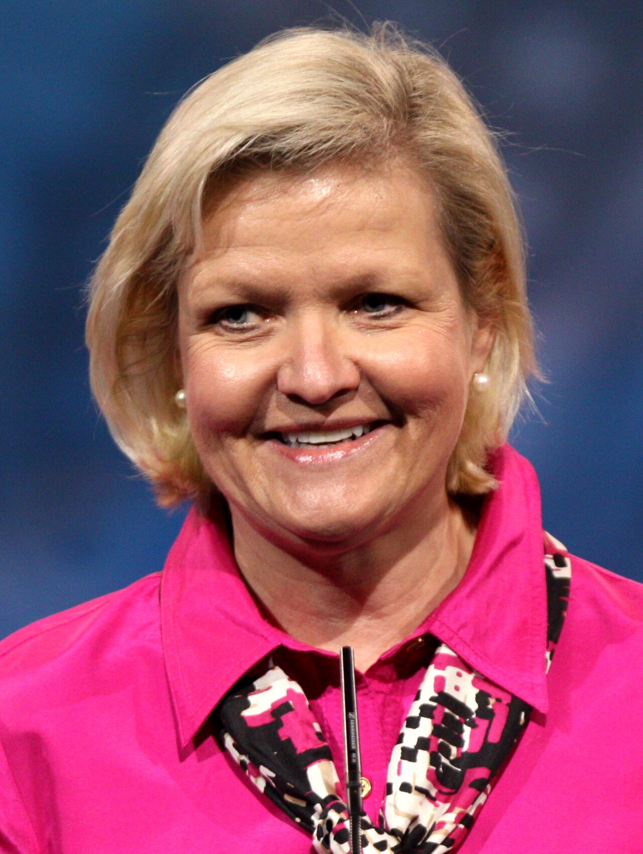 A woman with short blonde hair, wearing a pink shirt with a black and white patterned collar, smiling and looking directly at the camera.