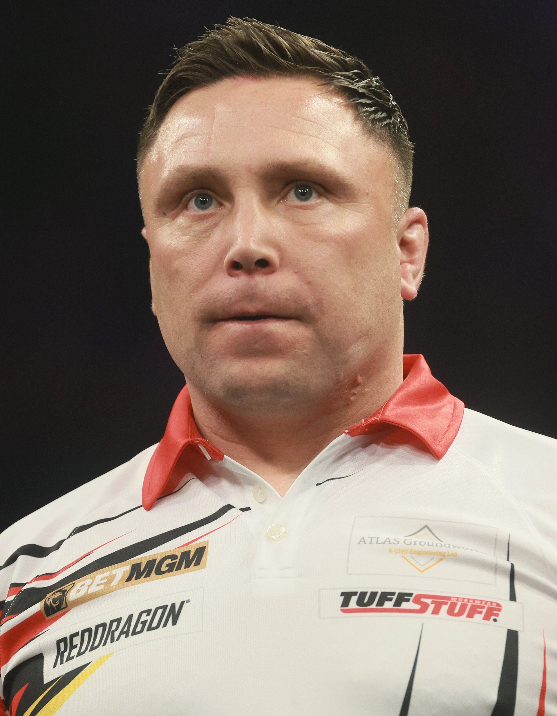 A man with short brown hair wearing a white polo shirt with red collar and several logos on it