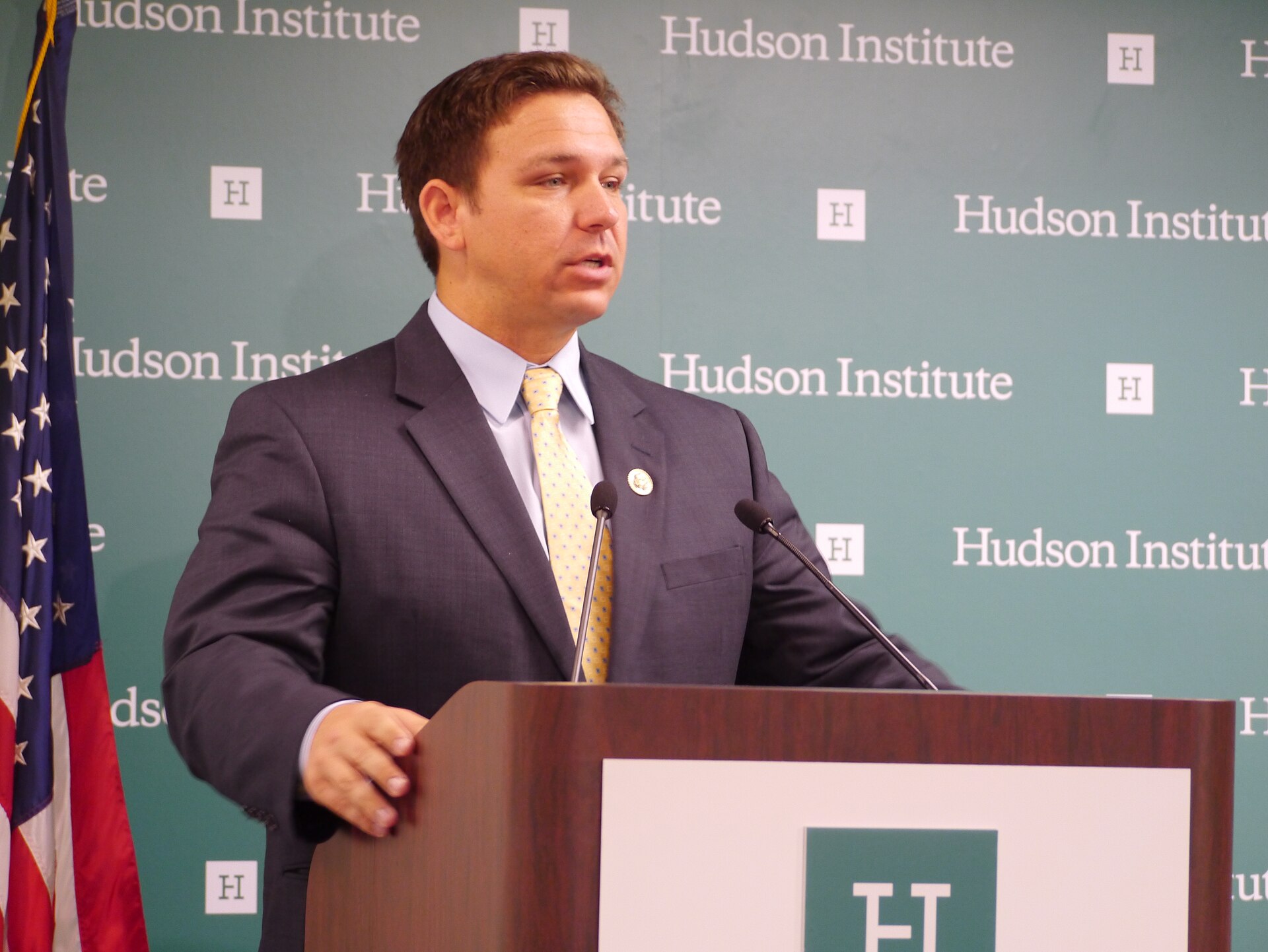 A man in a dark suit and yellow tie speaks at a podium, with an American flag to his left and a green backdrop with "Hudson Institute" repeated on it.