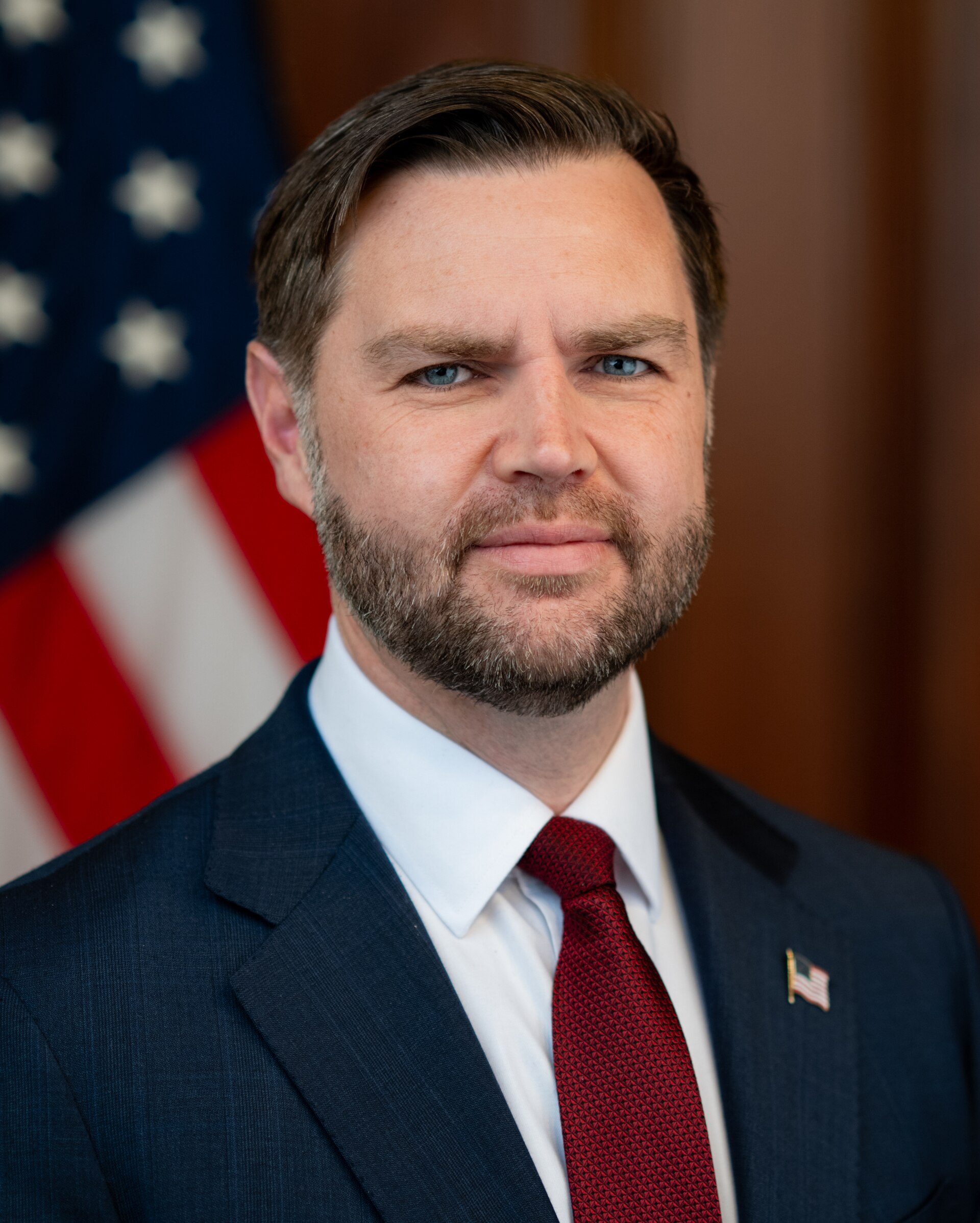 A man in a suit with a red tie and an American flag pin on his lapel, standing in front of a blurred American flag.