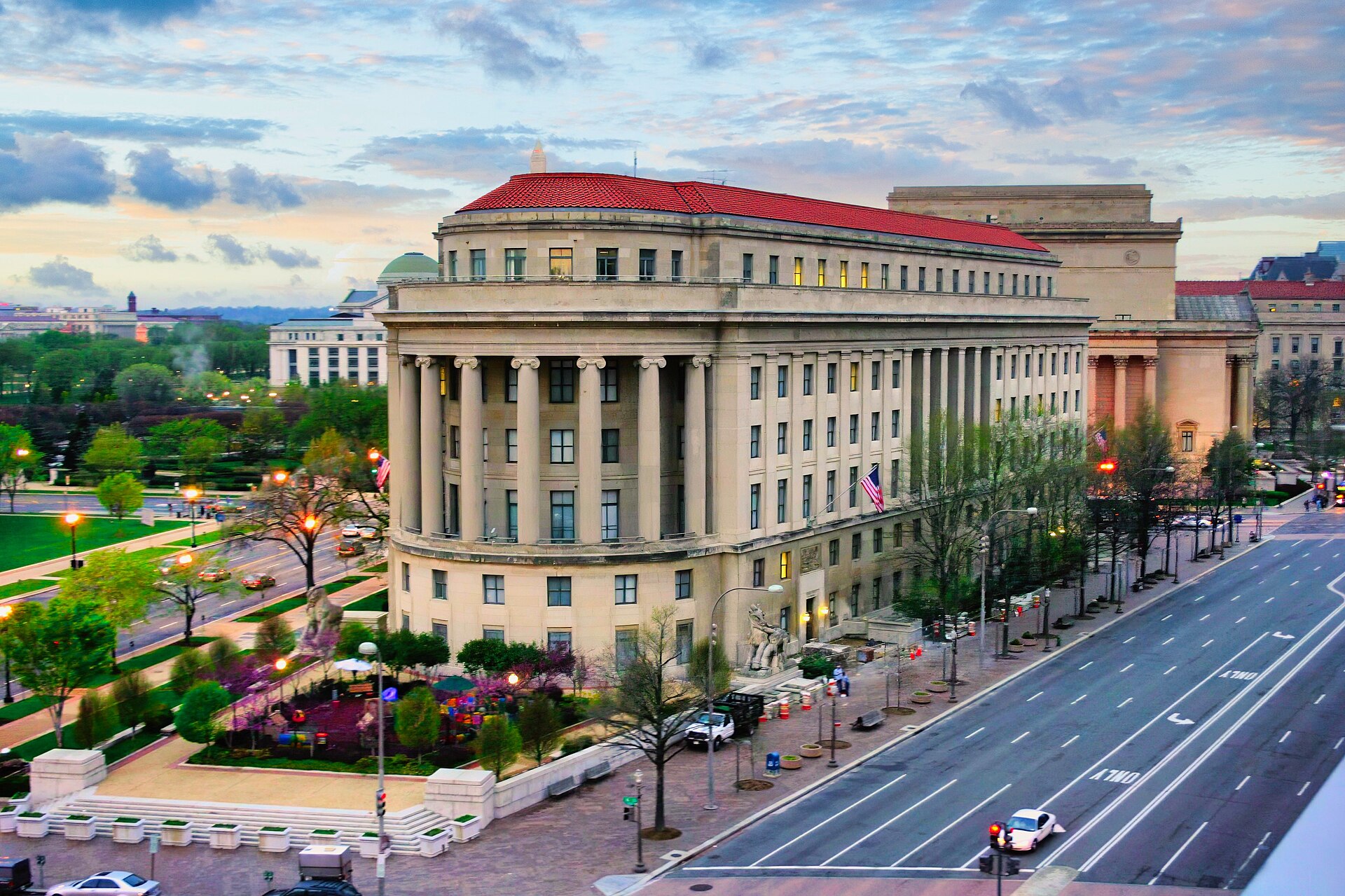 A large, classical-style building with a red roof and columns, seen from an elevated perspective at dusk. A street with cars runs alongside it, and a park area with trees and playground equipment is visible in front.