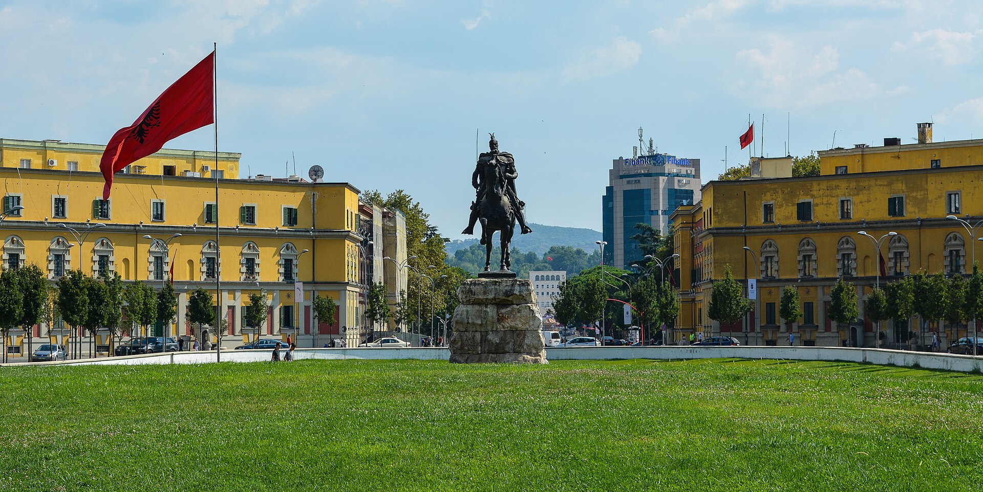 An equestrian statue stands in a grassy square in front of yellow buildings under a blue sky.