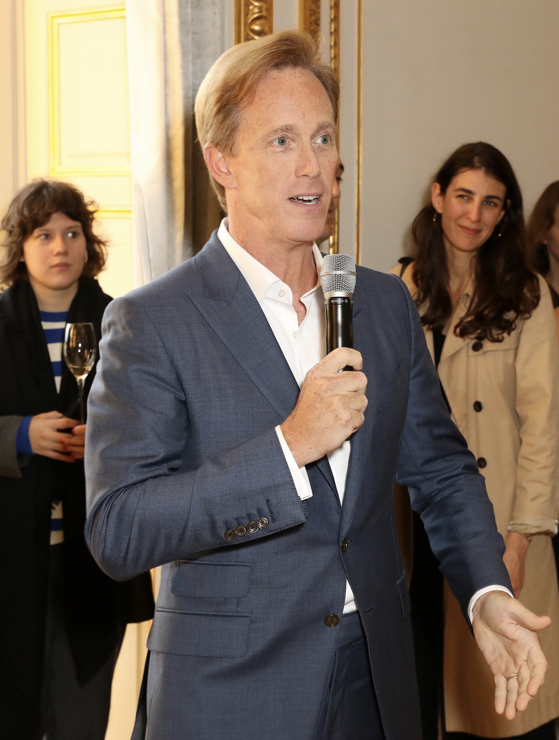 A middle-aged man with blond hair wearing a blue suit and white shirt holds a microphone and gestures with one hand.
