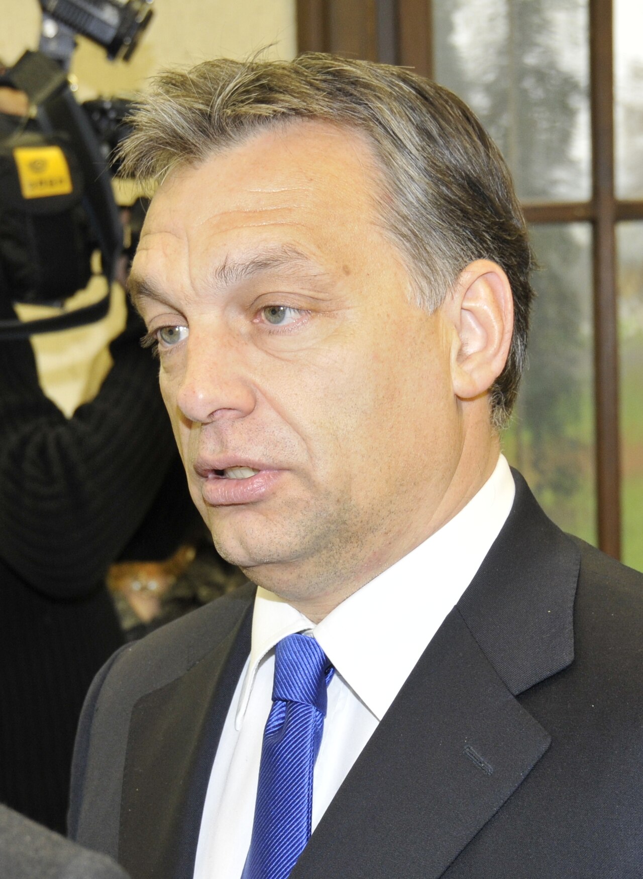 Viktor Orban, a middle-aged man with short graying hair, wearing a black suit, white shirt, and blue tie, looking to the left.