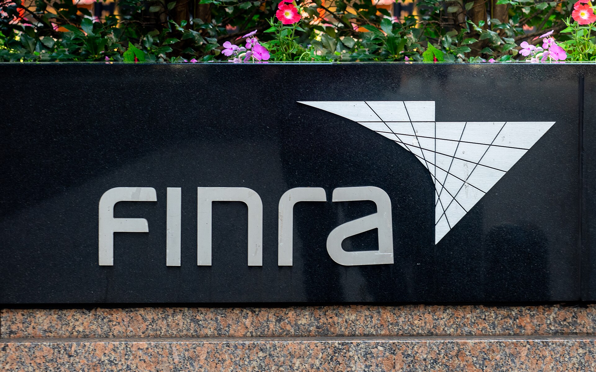 The FINRA logo is displayed in silver lettering on a black granite sign, with a stylized white triangle graphic to the right of the text. Green foliage and pink flowers are visible above the sign.