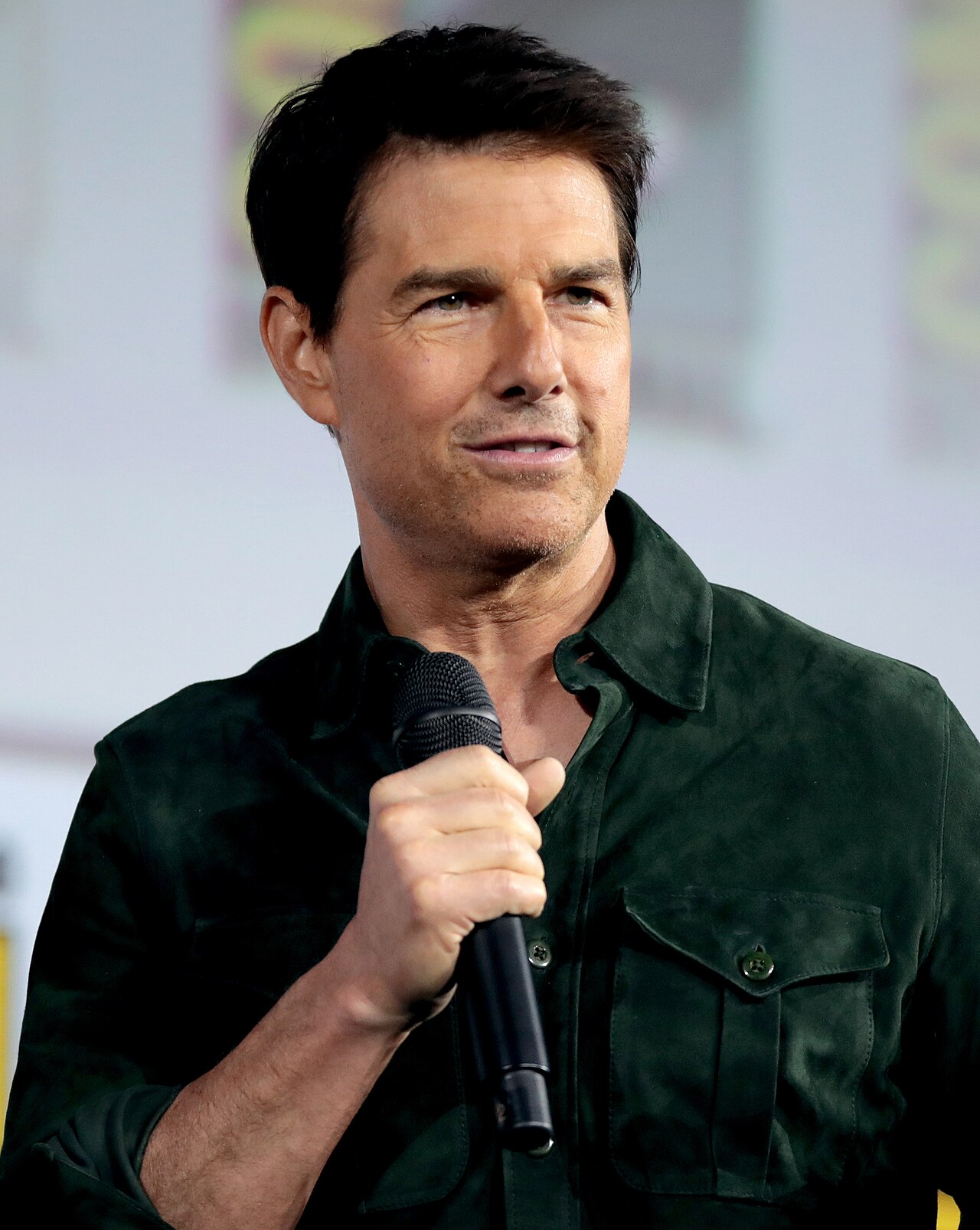Tom Cruise, wearing a dark green shirt, holding a microphone in his right hand and looking to the right with a slight smile