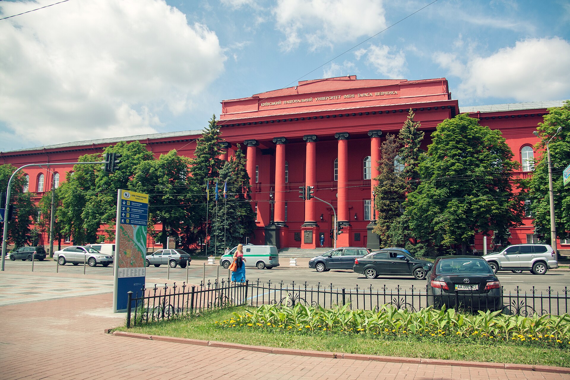 A large, red building with columns and the inscription "КИЇВСЬКИЙ НАЦІОНАЛЬНИЙ УНІВЕРСИТЕТ ІМЕНІ ТАРАСА ШЕВЧЕНКА" (Kyiv National University named after Taras Shevchenko) is visible under a cloudy sky. Cars are driving on the street in front of the building, and trees line the sidewalk.