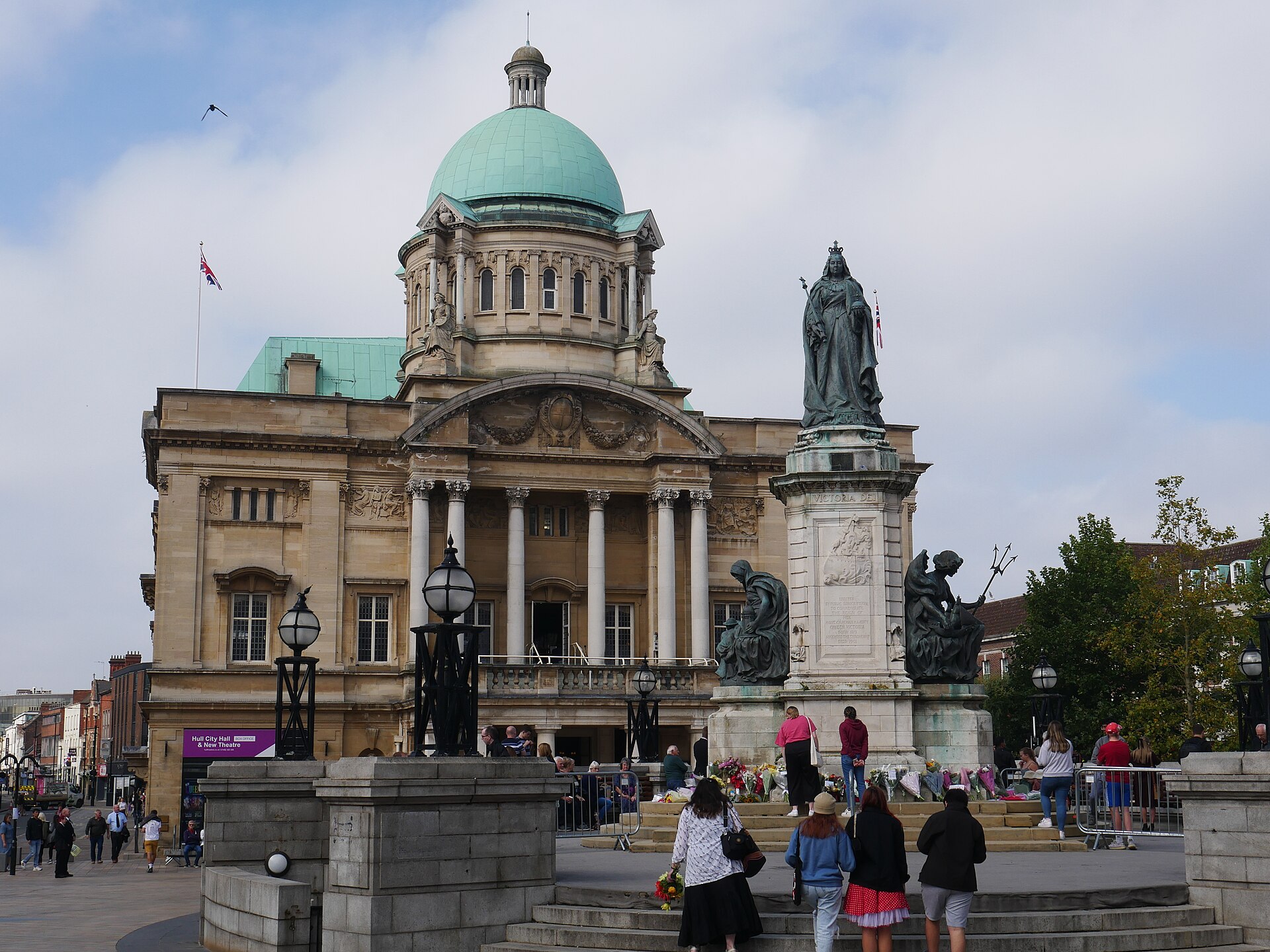 A statue of Queen Victoria stands in front of a domed building, with people gathered around the base of the statue.
