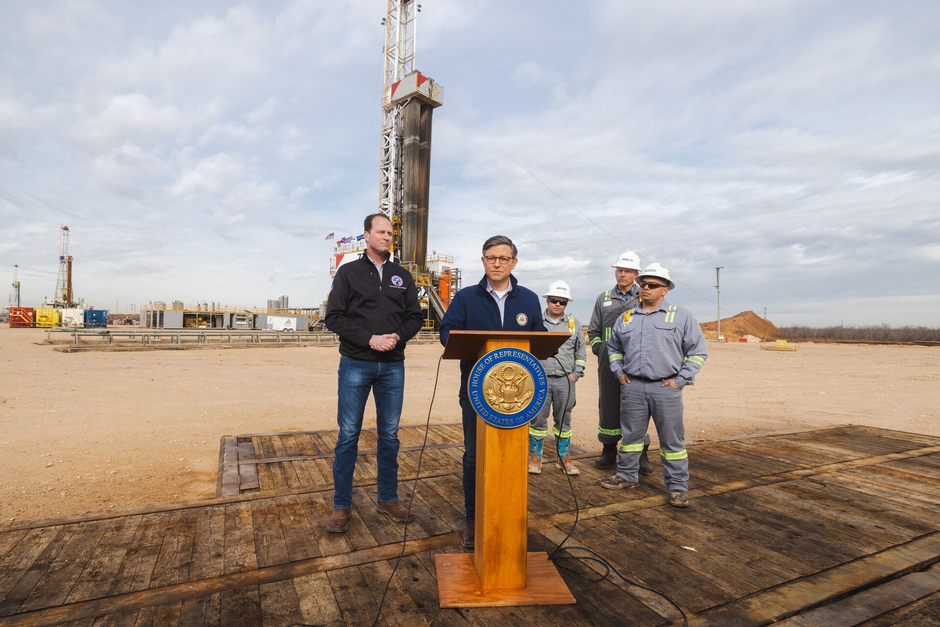 A man in a blue jacket stands at a podium with the "House of Representatives" seal, addressing a group of people in work clothes in front of a drilling rig.