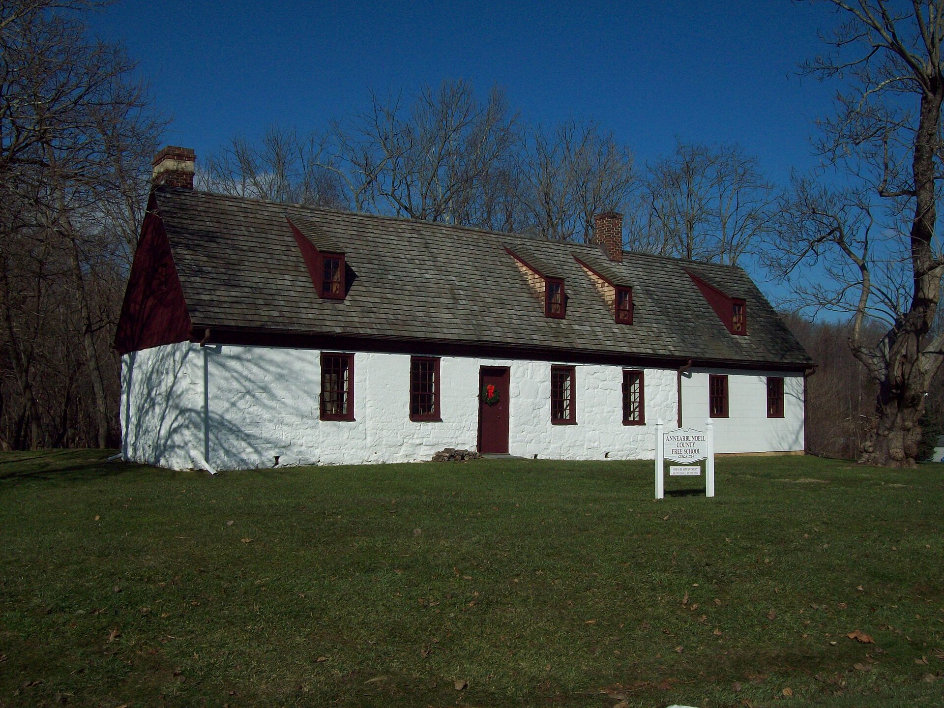 A white stone building with a dark roof and red trim, featuring multiple windows and dormers, stands on a grassy hill under a clear blue sky. A sign in front reads "ANNE ARUNDELL COUNTY FREE SCHOOL SINCE 1729".