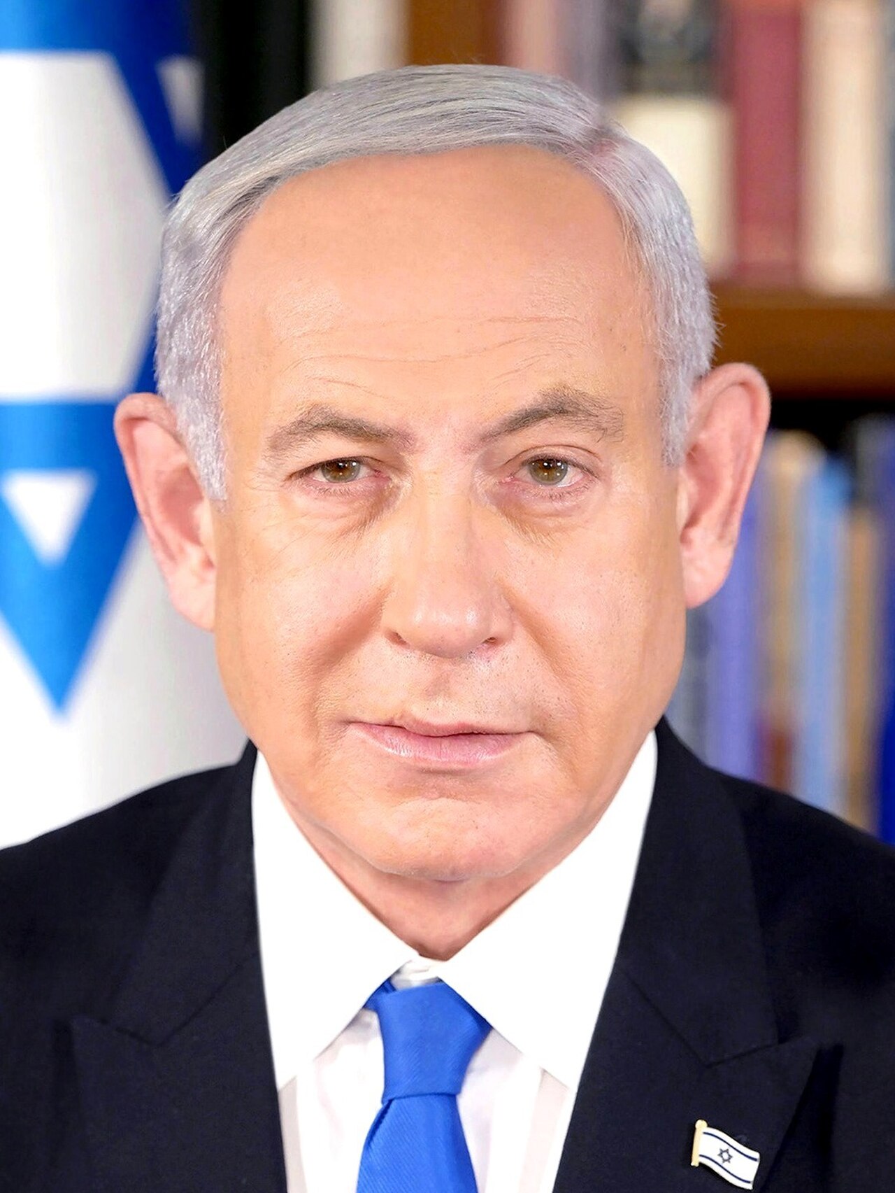Benjamin Netanyahu, wearing a suit, white shirt, blue tie, and an Israeli flag pin, looking directly at the camera.