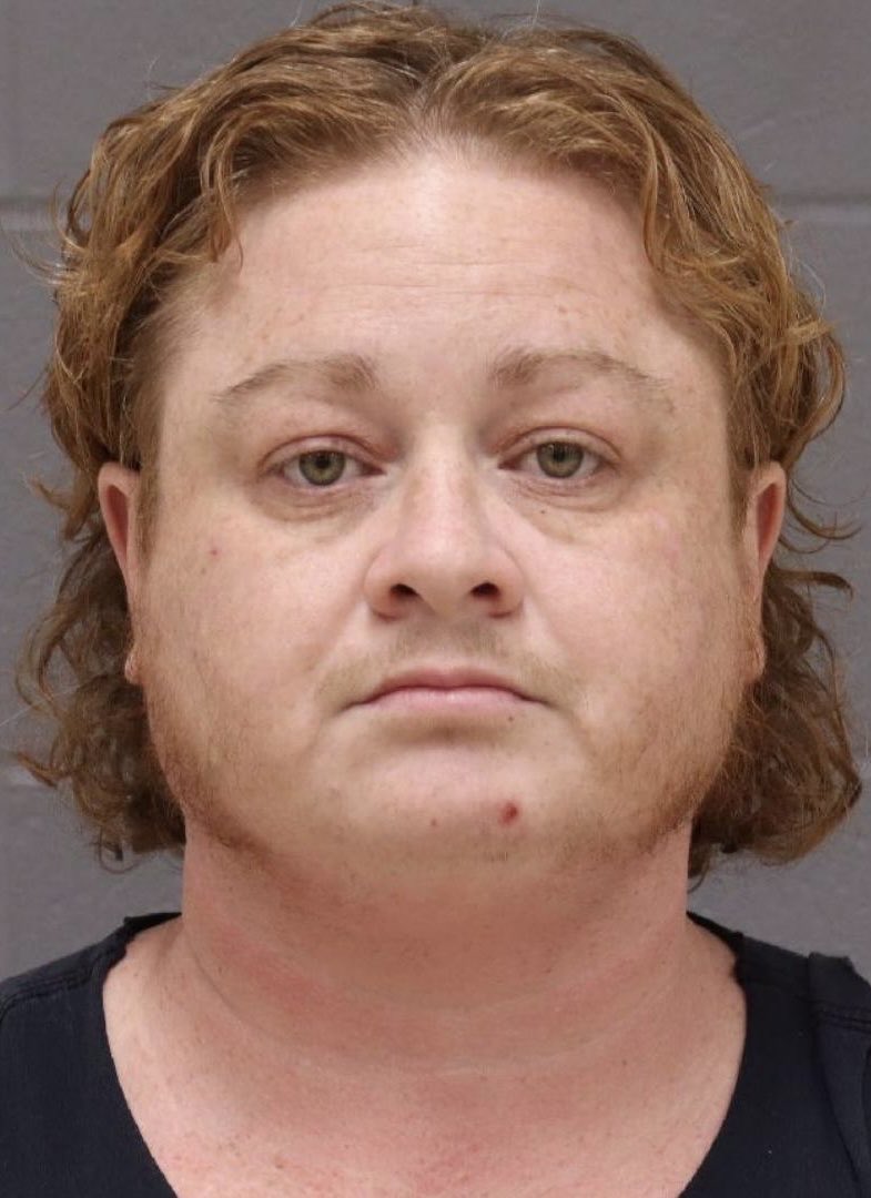 Booking photograph of Kieran Ansley, a person with shoulder-length reddish hair in a black tank top, against a cinder-block wall.