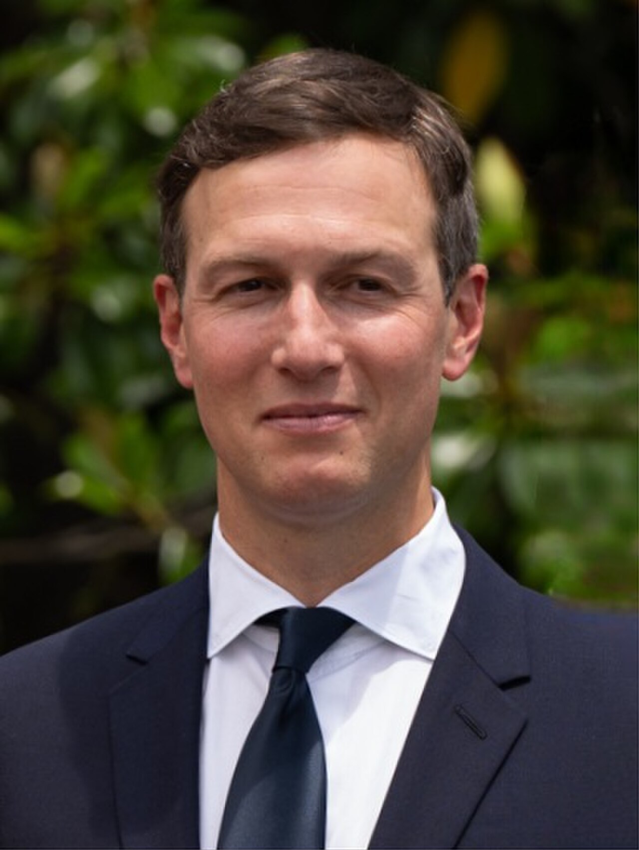 Jared Kushner, wearing a navy suit, white shirt, and navy tie, standing in front of greenery